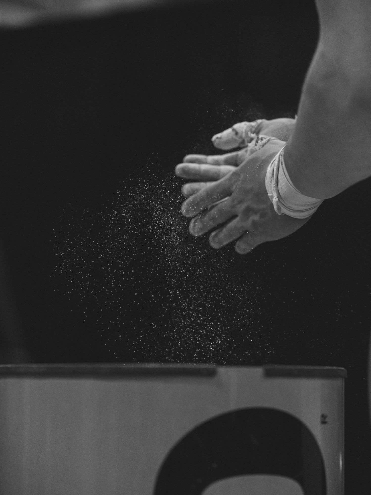 Black and white photo of a pair of hands chalking up above a chalk bowl. Chalk flakes are visible in a cloud below their hands.