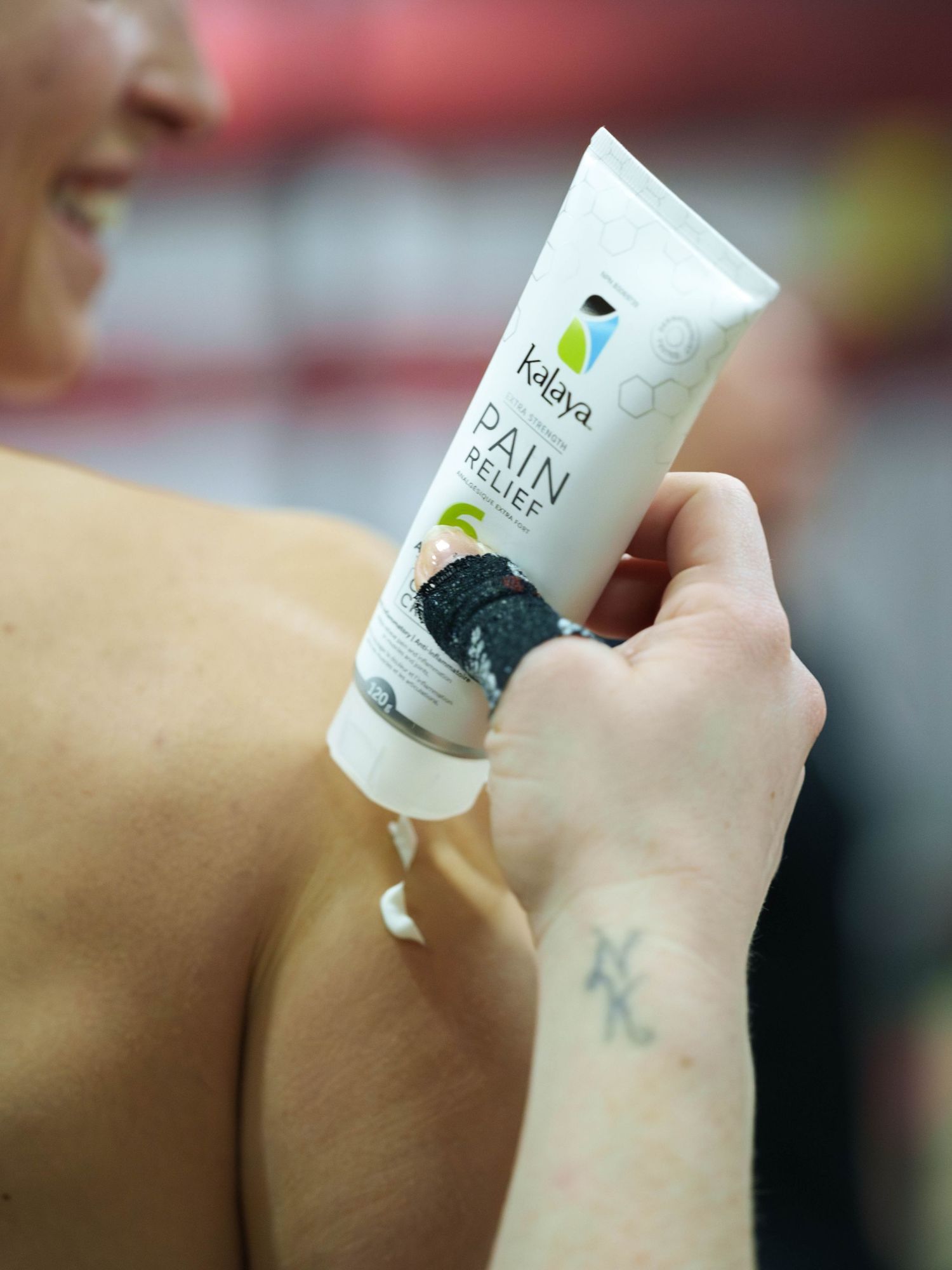Product photography for a tube of pain relief cream. An anonymous woman applies the cream to the shoulder of a female athlete.