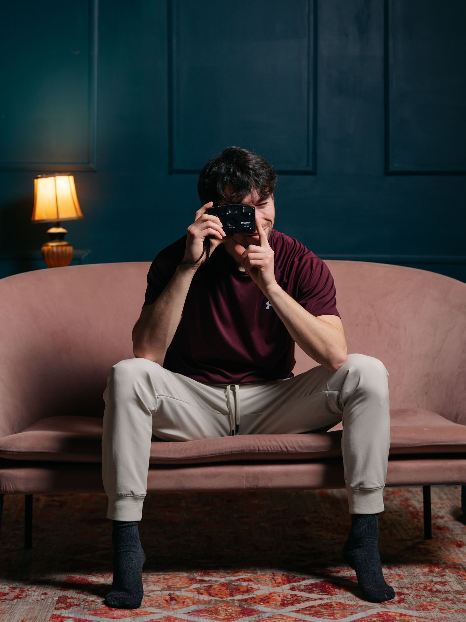 Flash studio portrait of a young white man sitting on a pink couch in front of a green wall holding a camera prop up to his eyes.