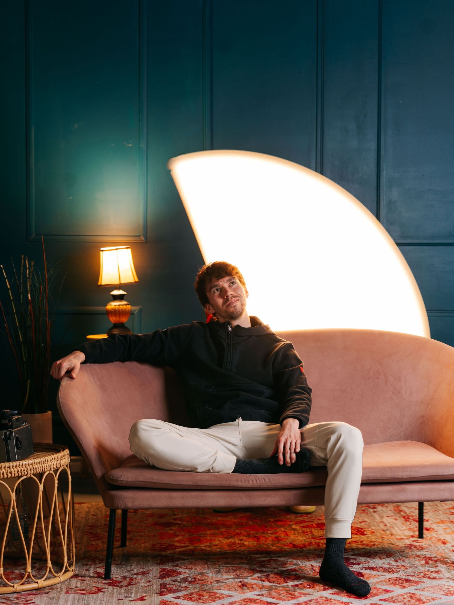 Flash studio portrait of a young white man sitting on a pink couch in front of a green wall with a windmill tube light motion blur behind.