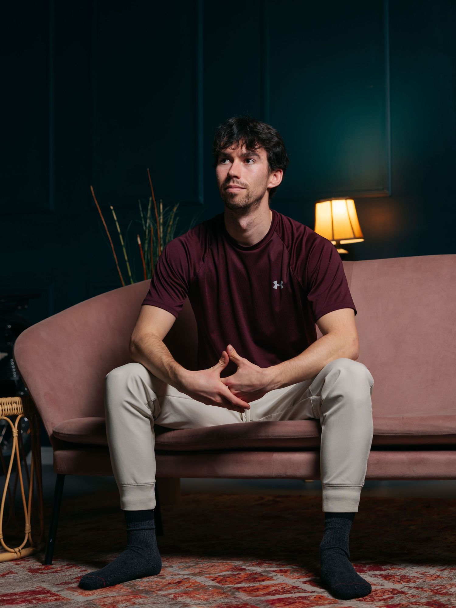 Flash studio portrait of a young white man sitting on a pink couch in front of a green wall looking off into the distance in contemplation.