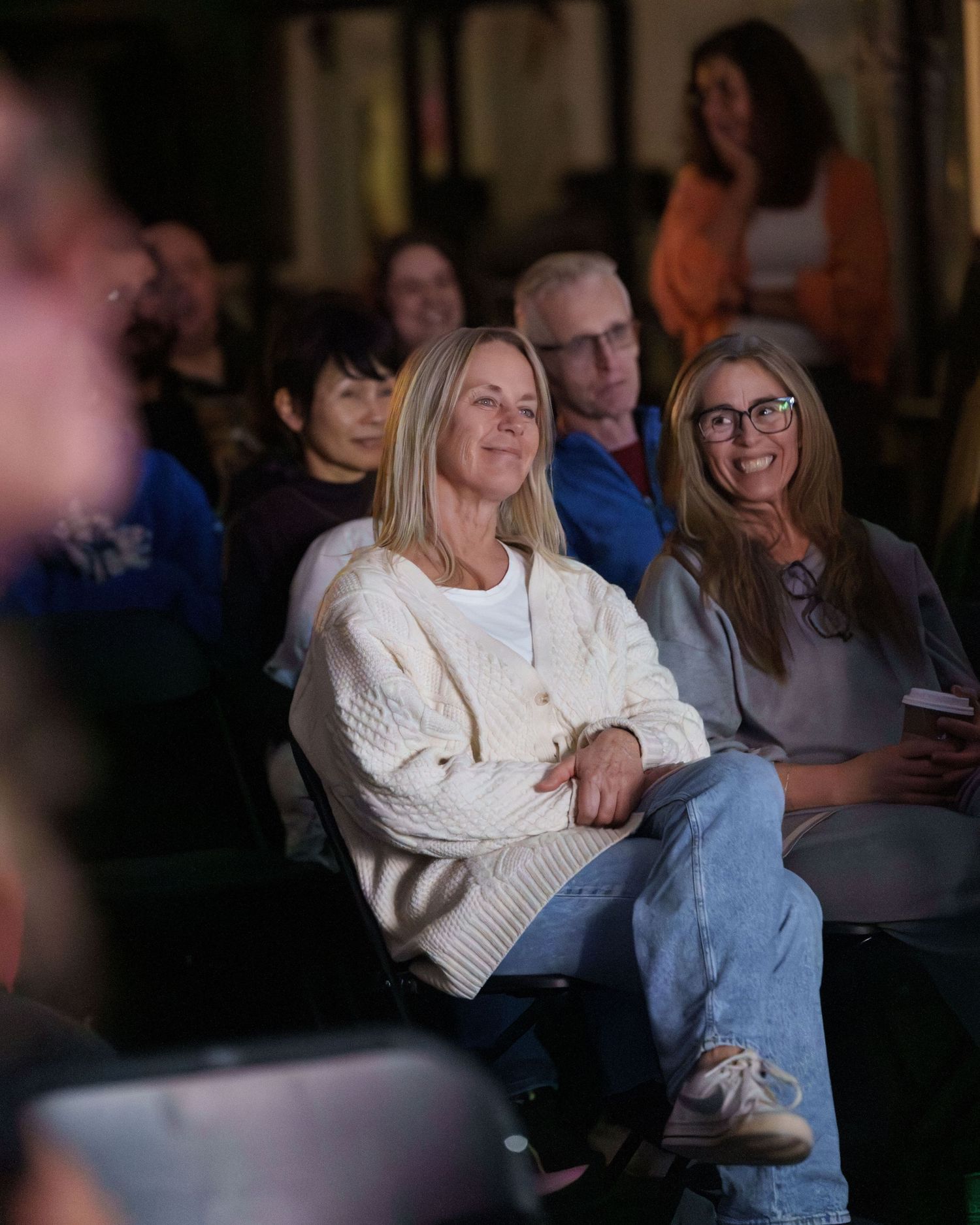 Two white, middle age women watching a documentary about their friend. One smiles while watching the screen. The other smiles looking at her