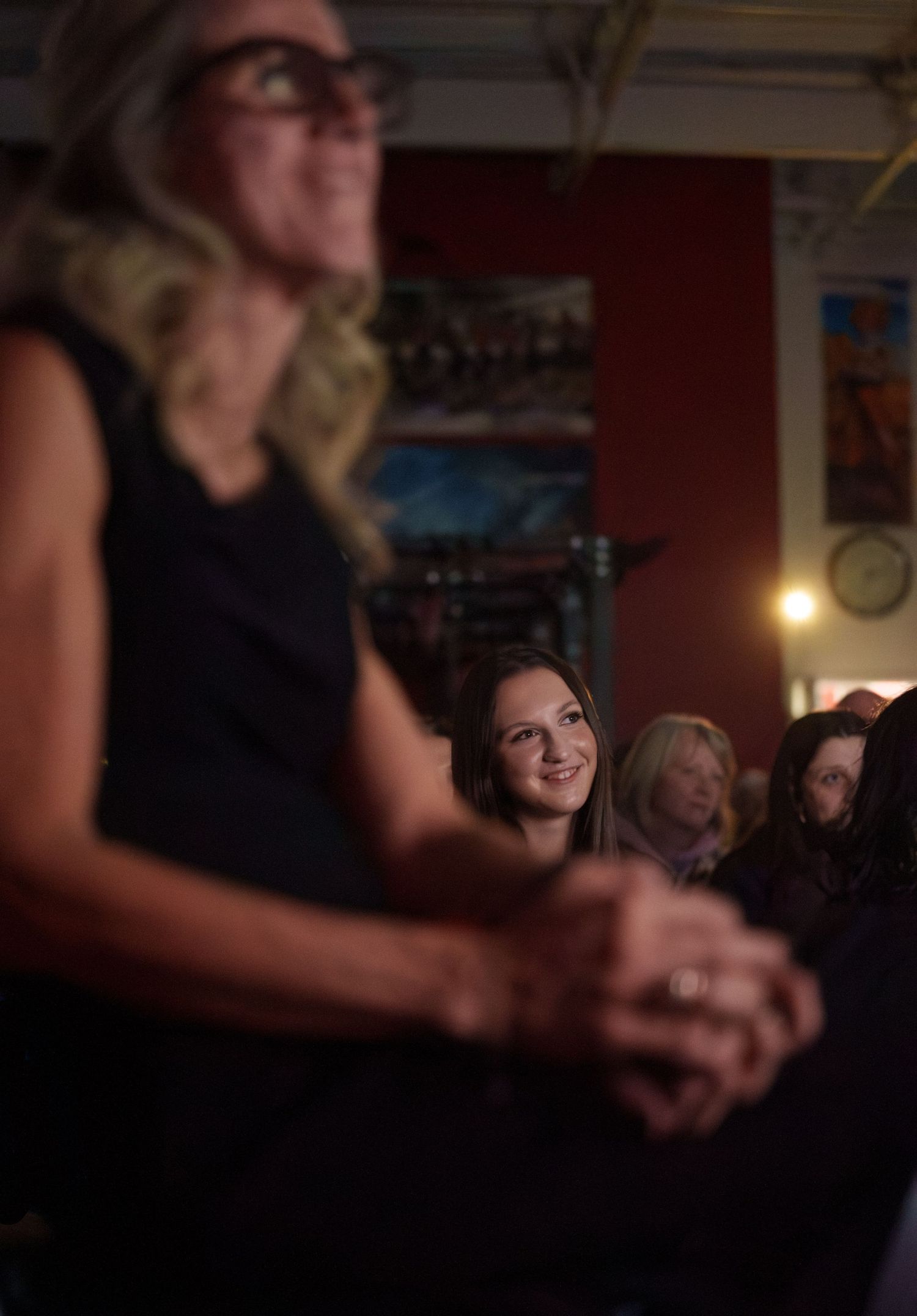 Two white women. A middle aged mom blurry in the foreground. In the background, her daughter smiling while watching a film about her mom.