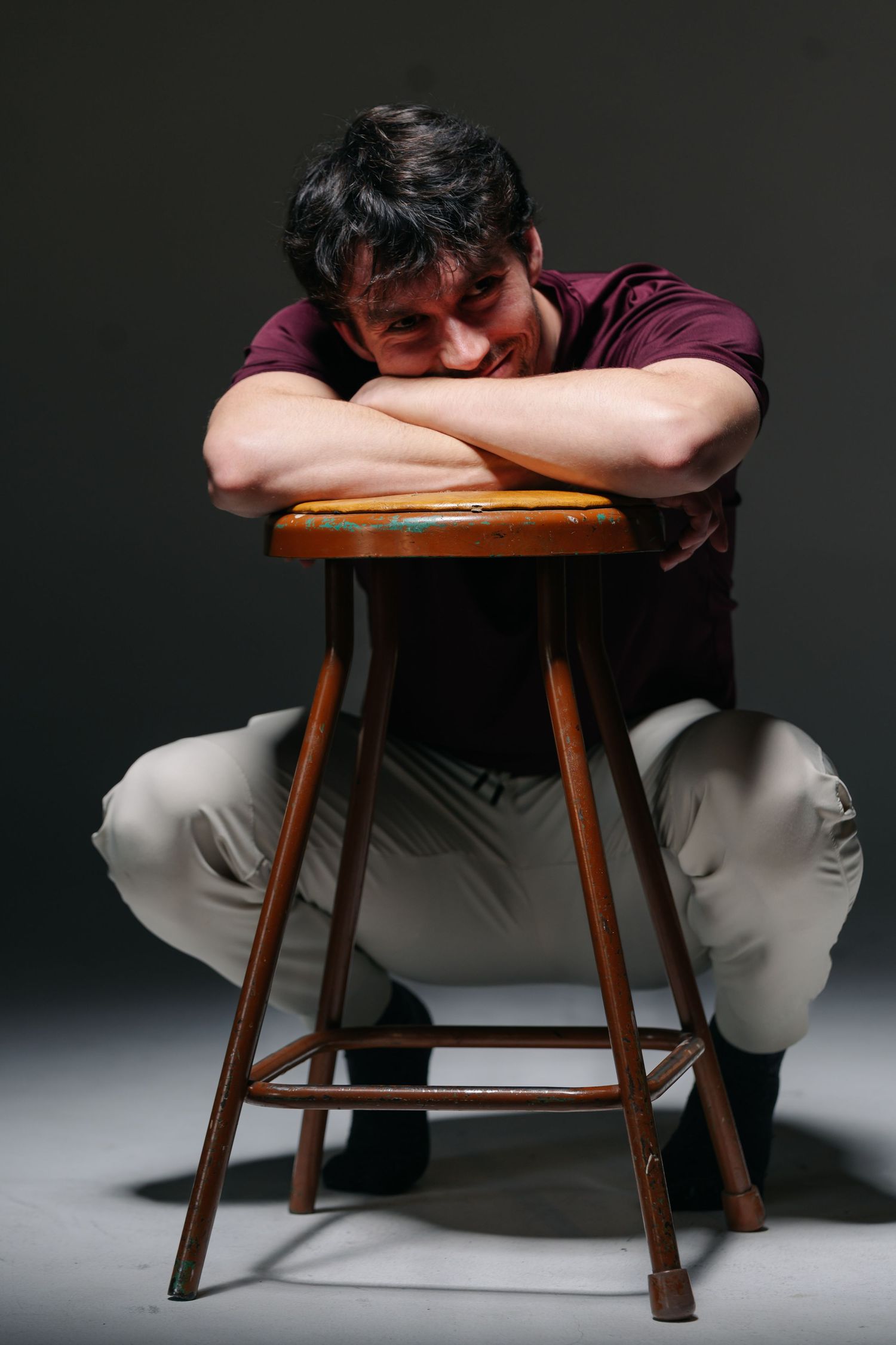 Young white man rests his head into his arms, which are crossed atop a stool as he crouches behind it. He smiles softly looking off camera.