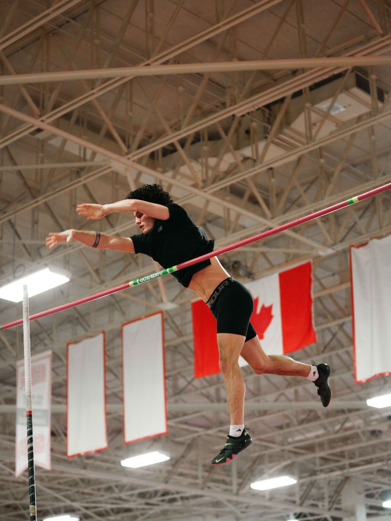 A young male polevaulter in dark clothing illuminated by the lights in the gym as he descends from a successful attempt above the bar.