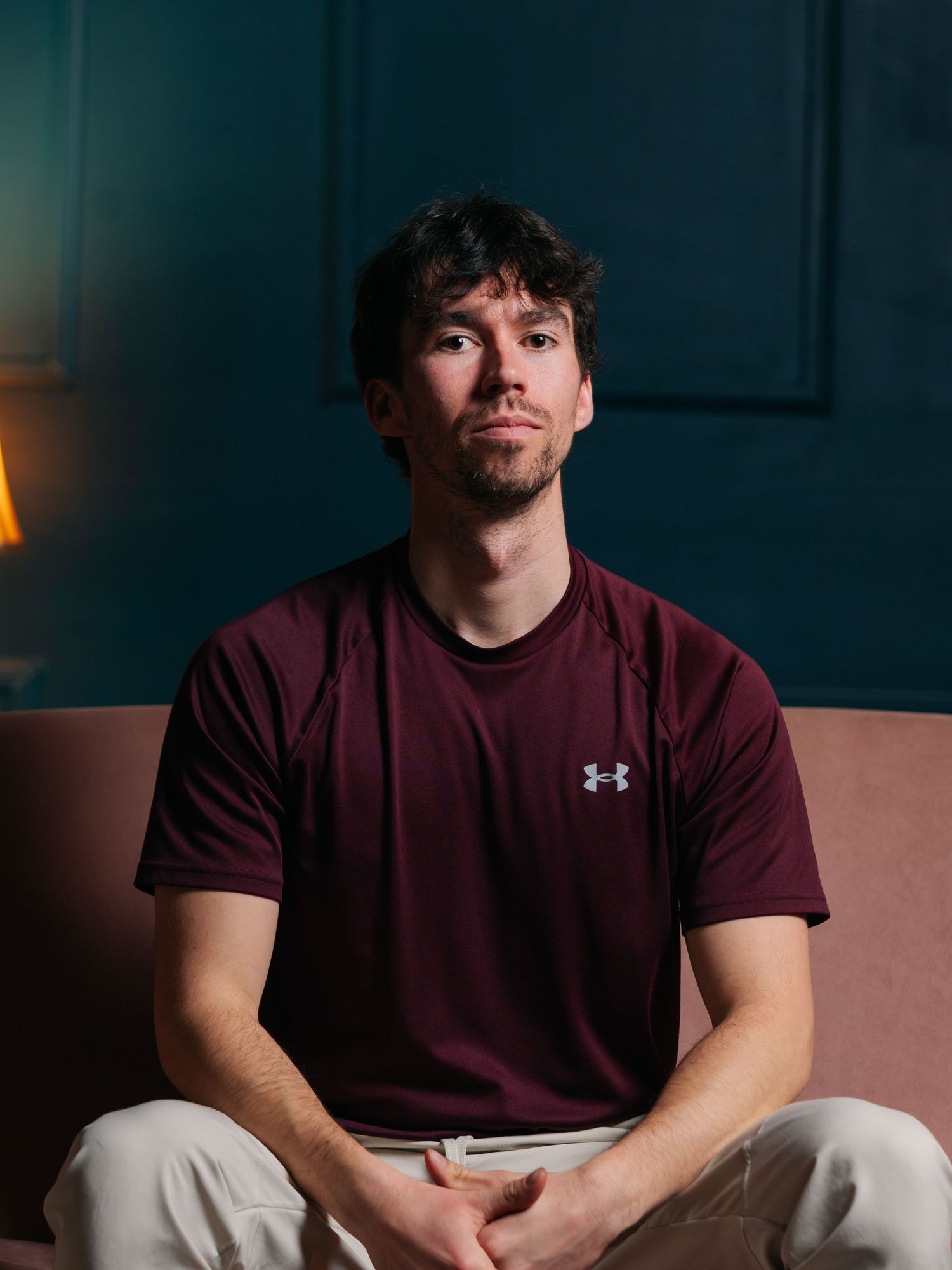 Flash studio portrait of a young white man sitting on a pink couch in front of a green wall looking down and serious at the camera.