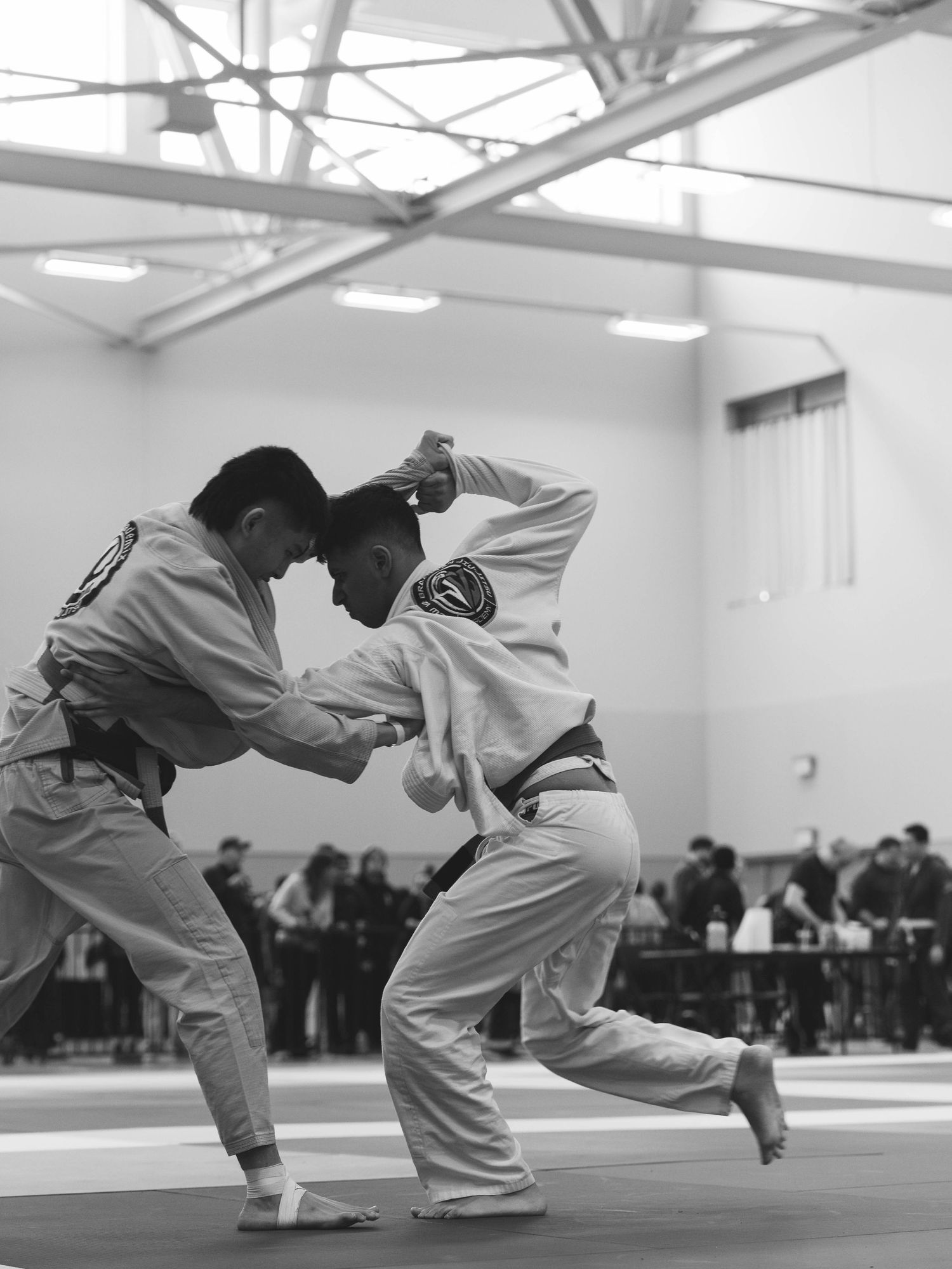 Black and white silhouette photo of two young male BJJ fighters in a large room lit with natural sunlight.