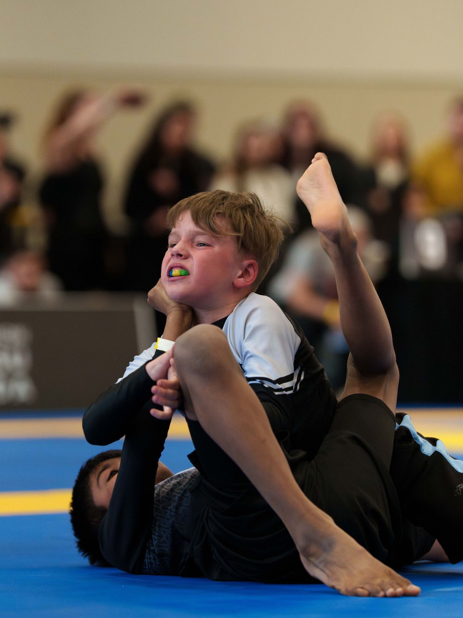 Two young boy BJJ fighters in rash guards grapple on the ground.