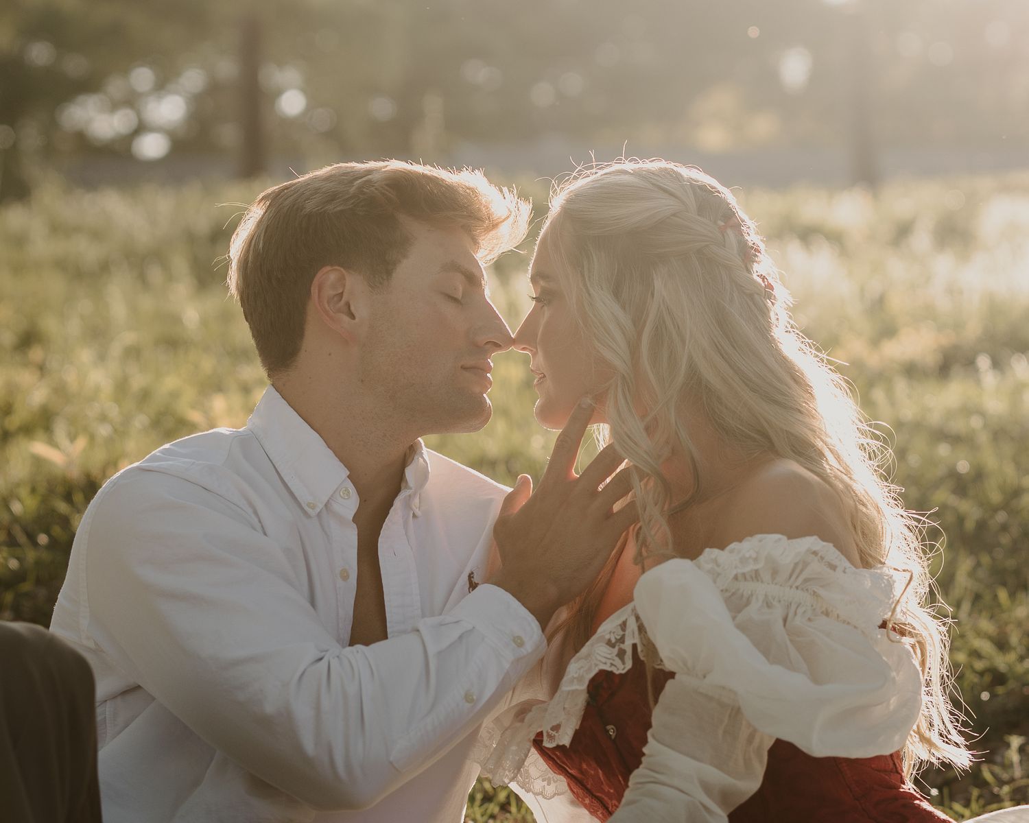 A romantic couple shares an intimate moment in a sunlit meadow, the woman in a white off-shoulder top and red skirt.
