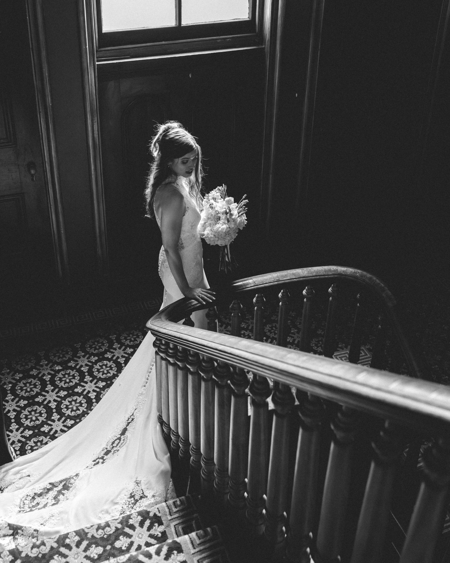 Black and white photo of a bride in a wedding gown holding a bouquet on an elegant staircase.