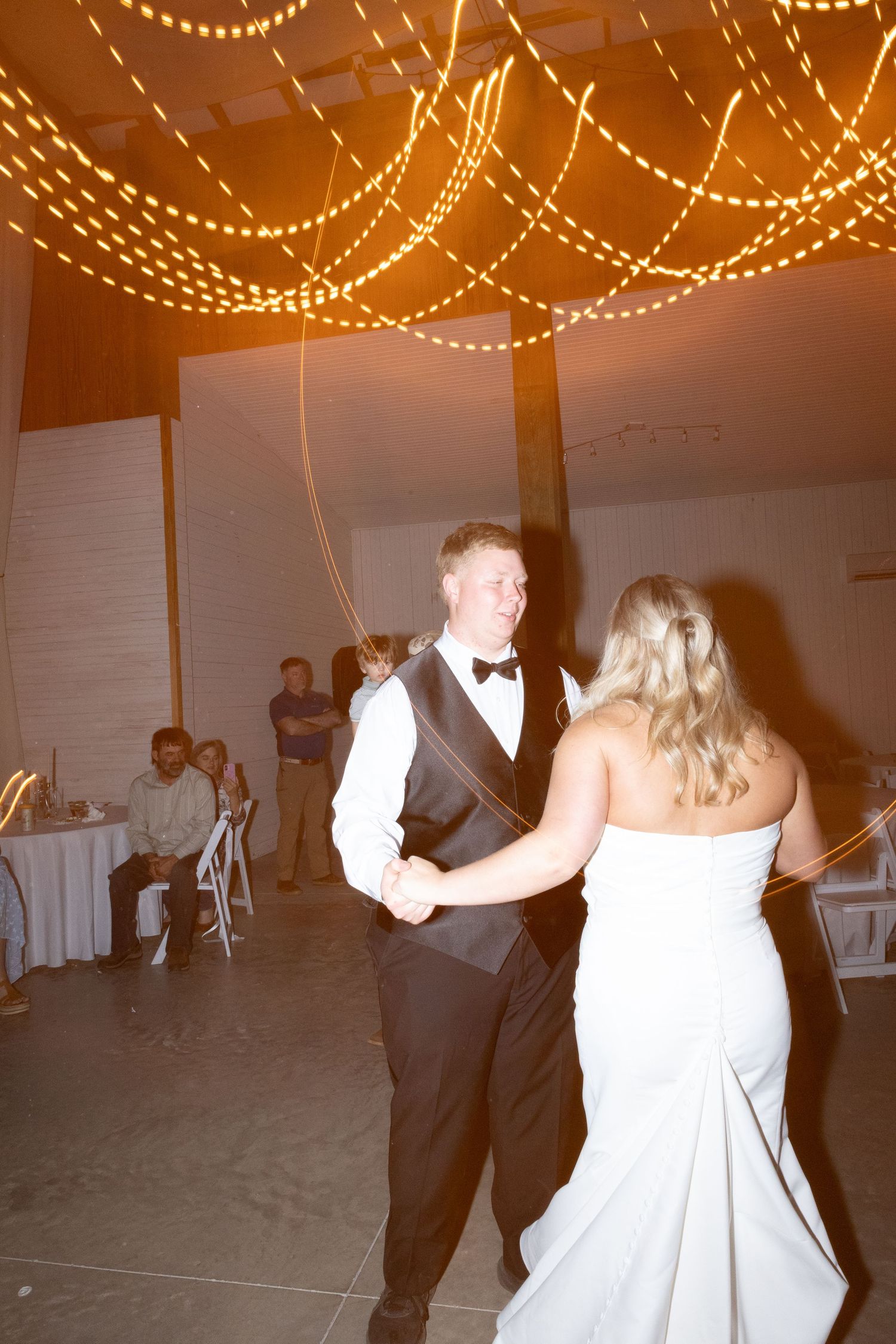 Bride and groom share their first dance under warm string lights at elegant wedding reception.