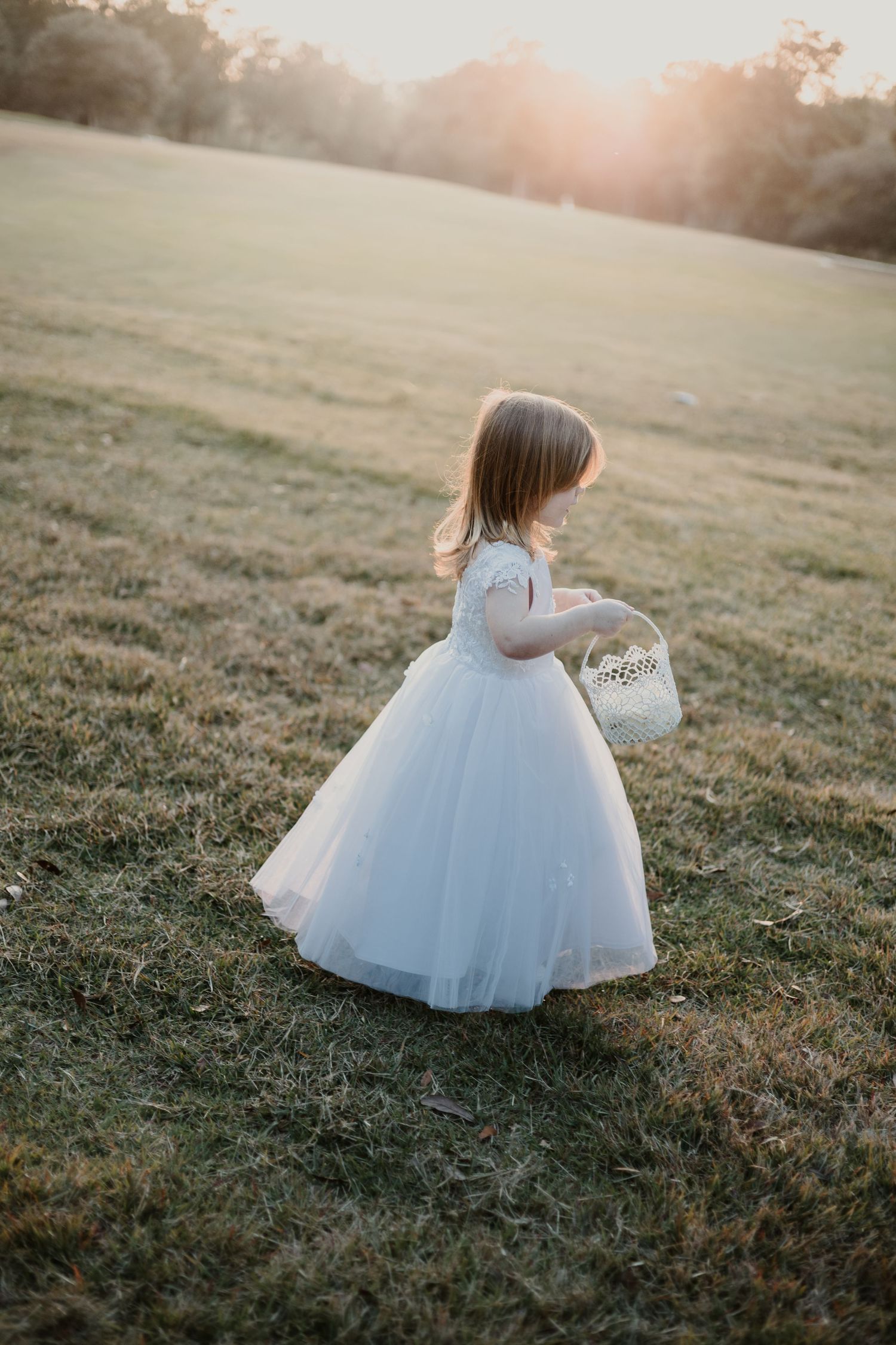 Young flower girl in white tulle dress holding basket walks through sunlit field at golden hour.