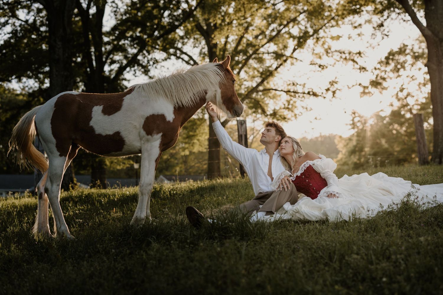Bride and groom sitting on grass petting a paint horse at golden hour during outdoor wedding photo session.