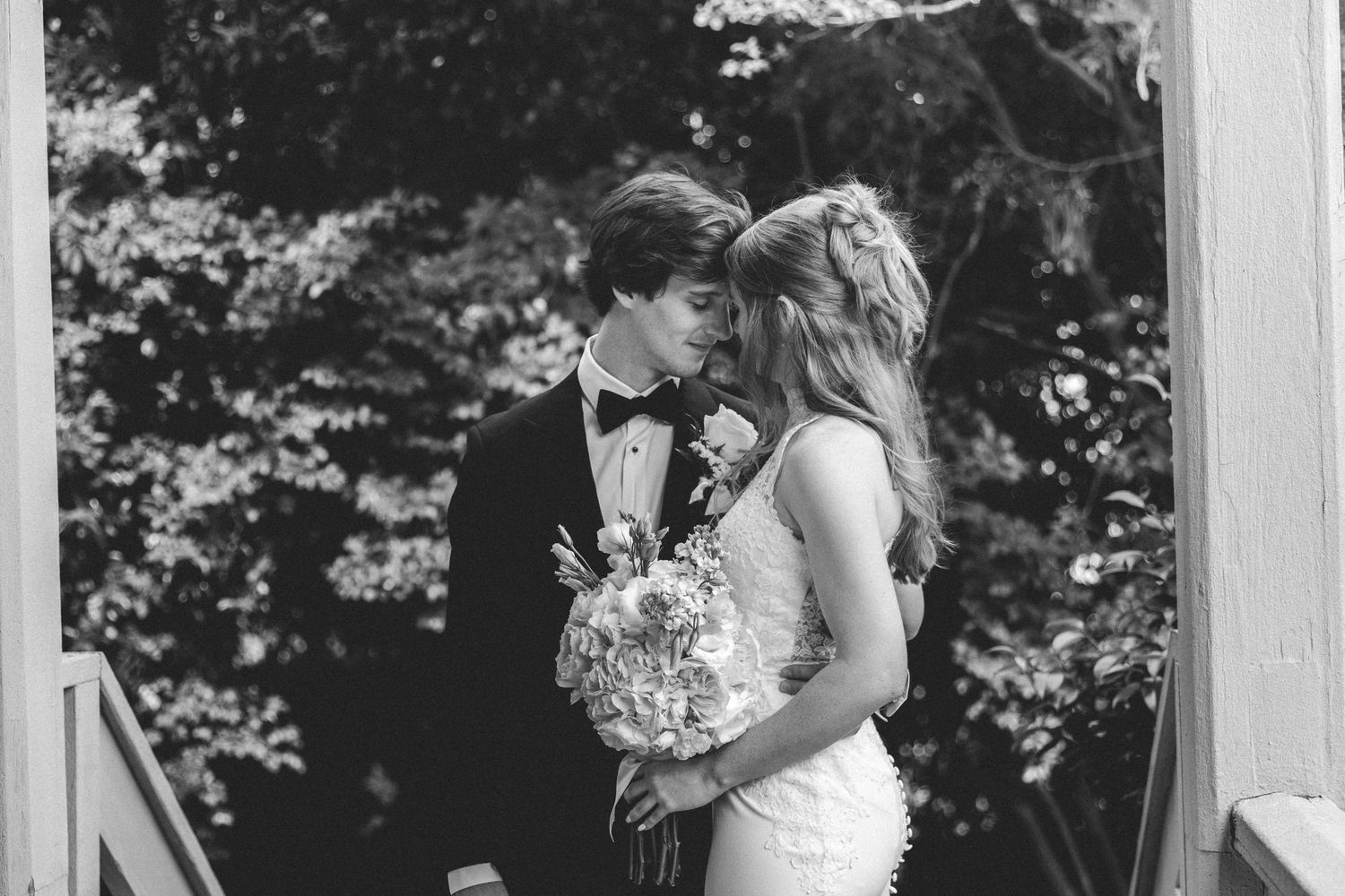 Black and white photo of a bride and groom sharing an intimate moment, bride holding a bouquet outdoors.