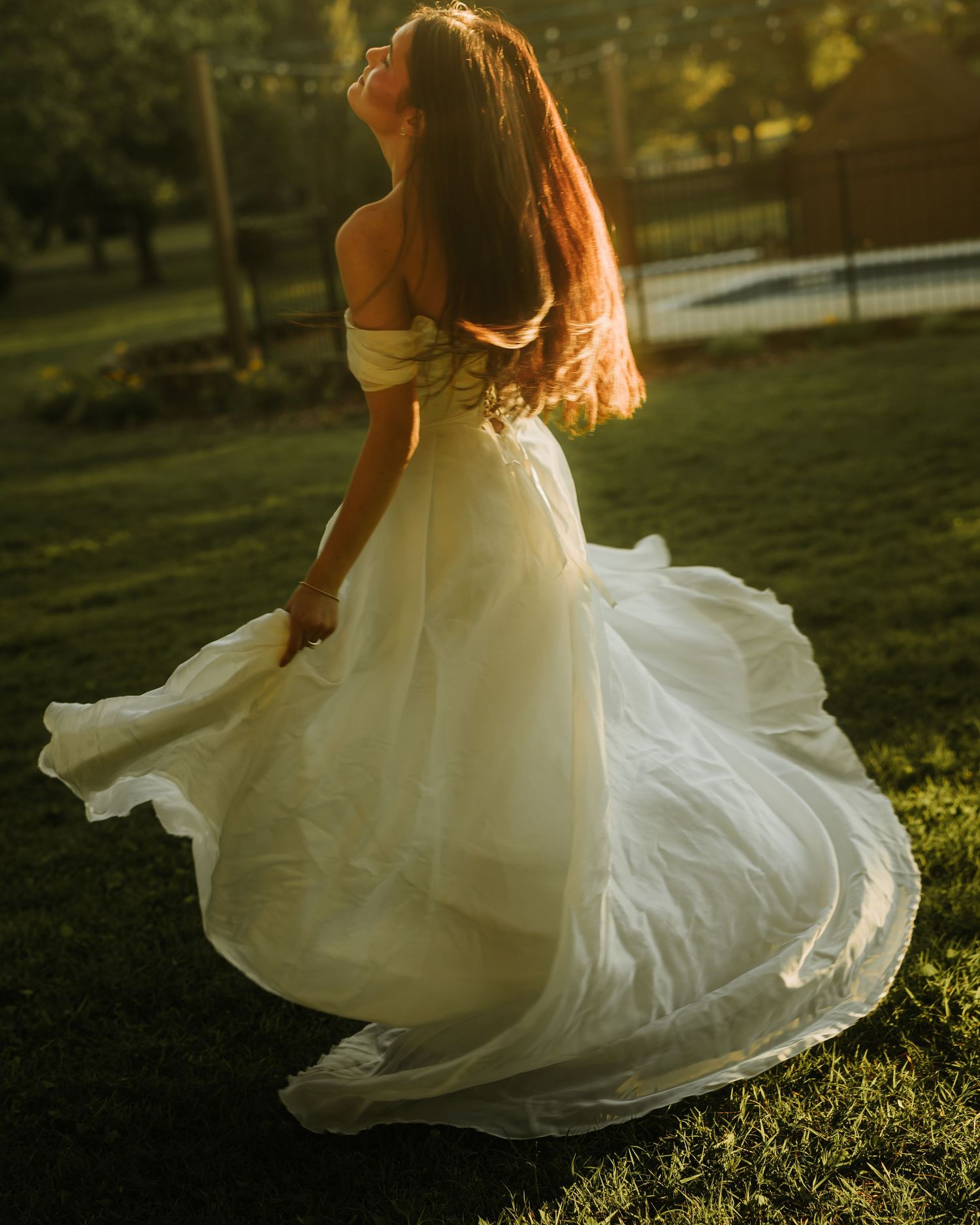 A bride in a flowing off-shoulder white gown twirls gracefully on green grass in golden sunset light.