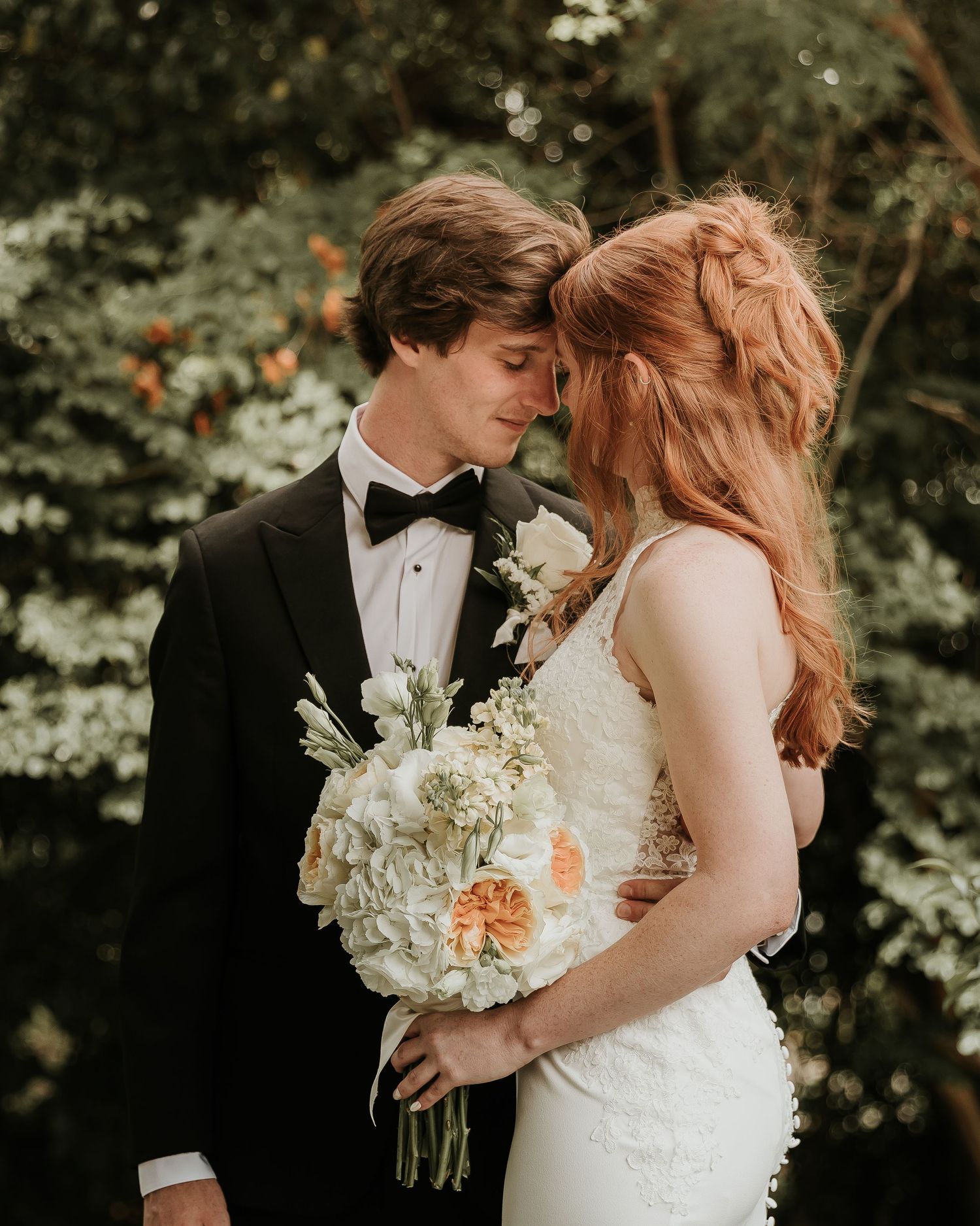 A bride and groom share a romantic forehead touch moment, she holds a white and peach bouquet, surrounded by lush greenery.