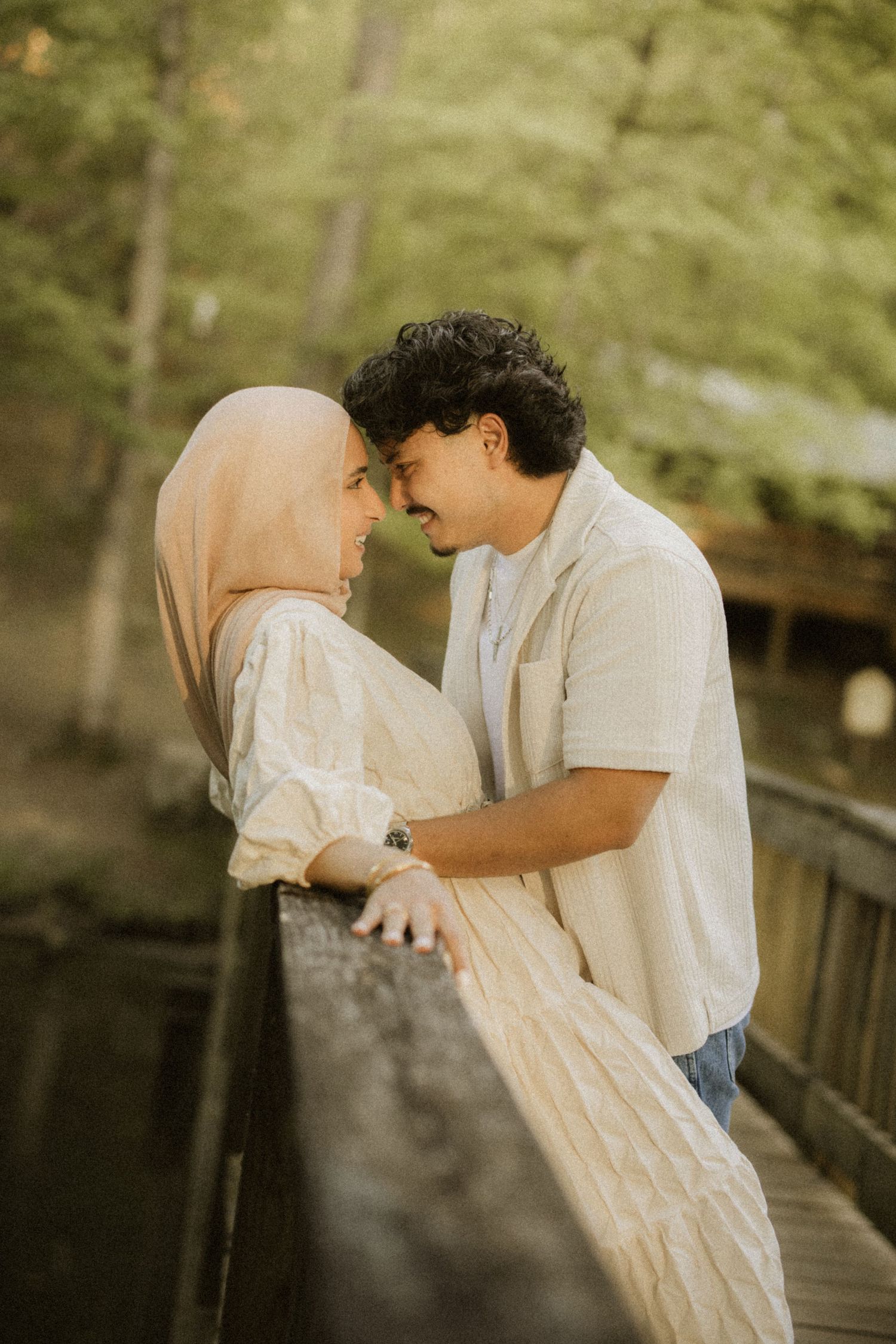 A romantic couple sharing an intimate moment on a wooden bridge surrounded by lush green trees in a park.