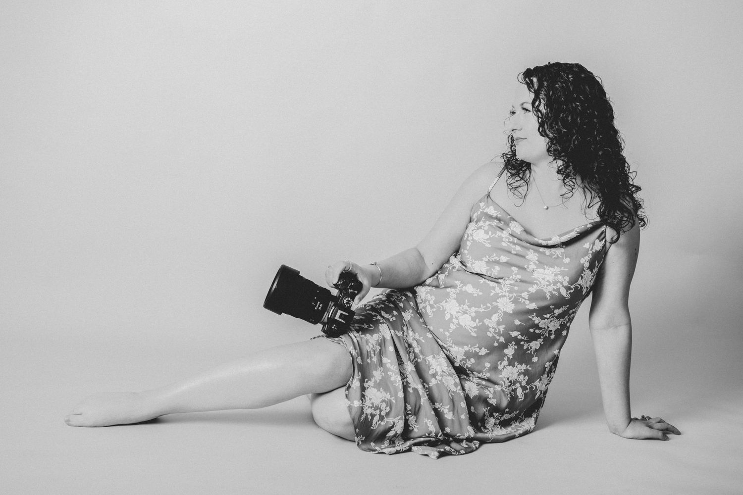 Black and white photo of a woman in a floral dress sitting on the floor holding a camera.
