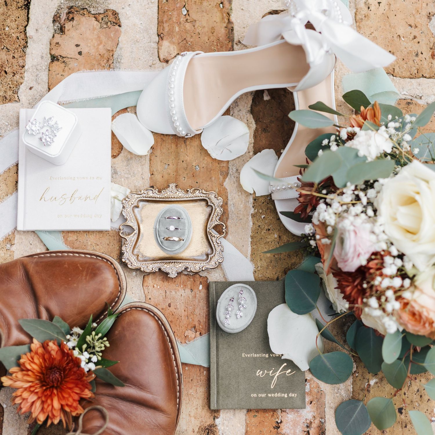 Elegant wedding flat lay with bridal heels, rings, bouquet, boutonniere, and vow books on rustic brick background.