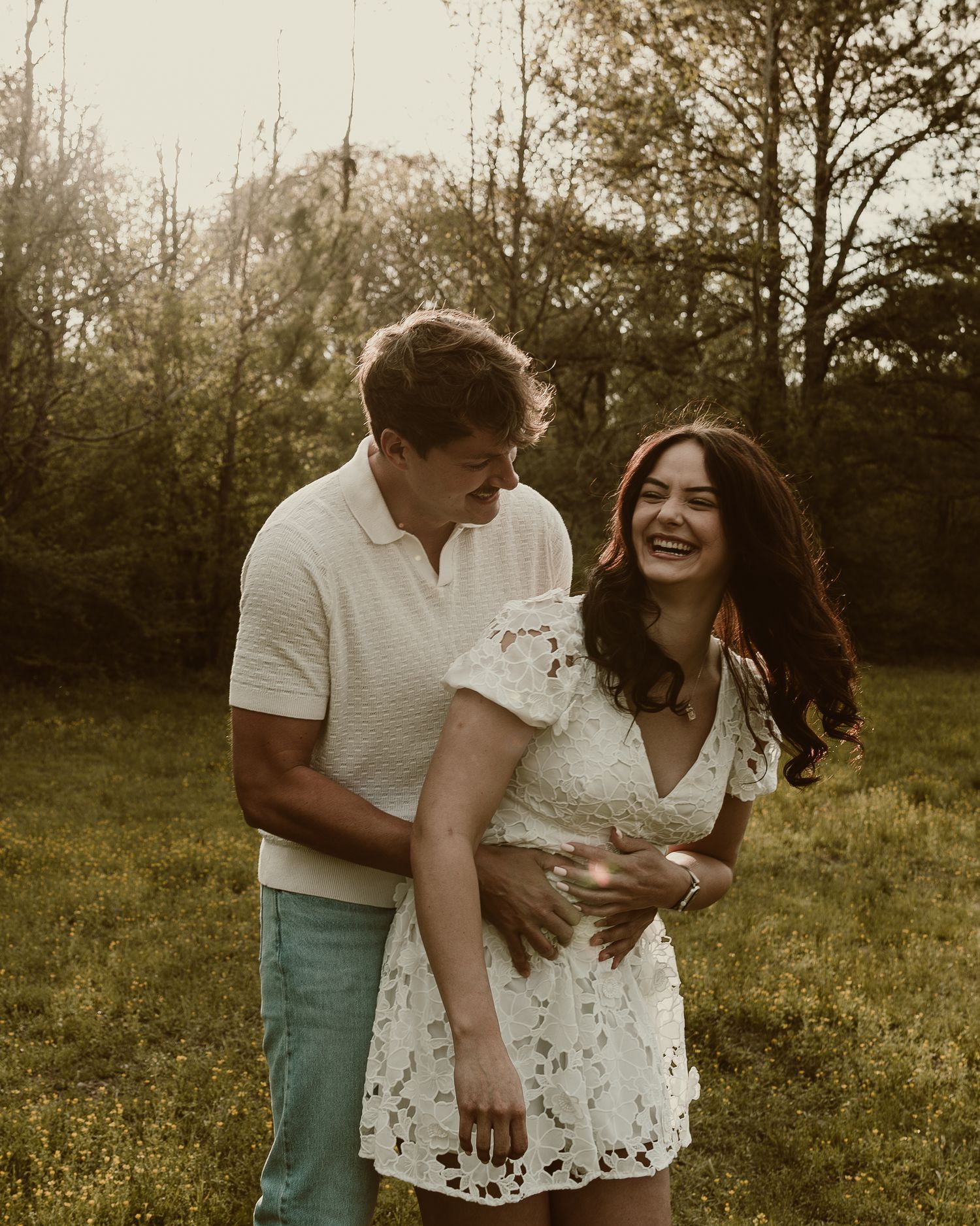 Happy couple laughing together in a sunlit forest clearing, woman in white lace dress.