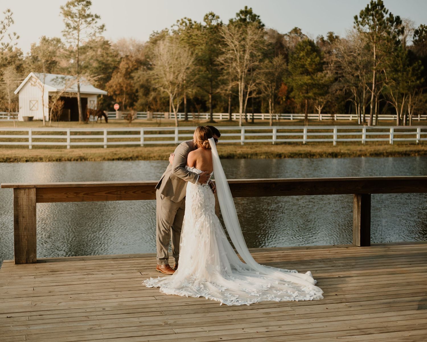 Bride and groom share a kiss on a wooden dock overlooking a pond at a rustic farm wedding venue.