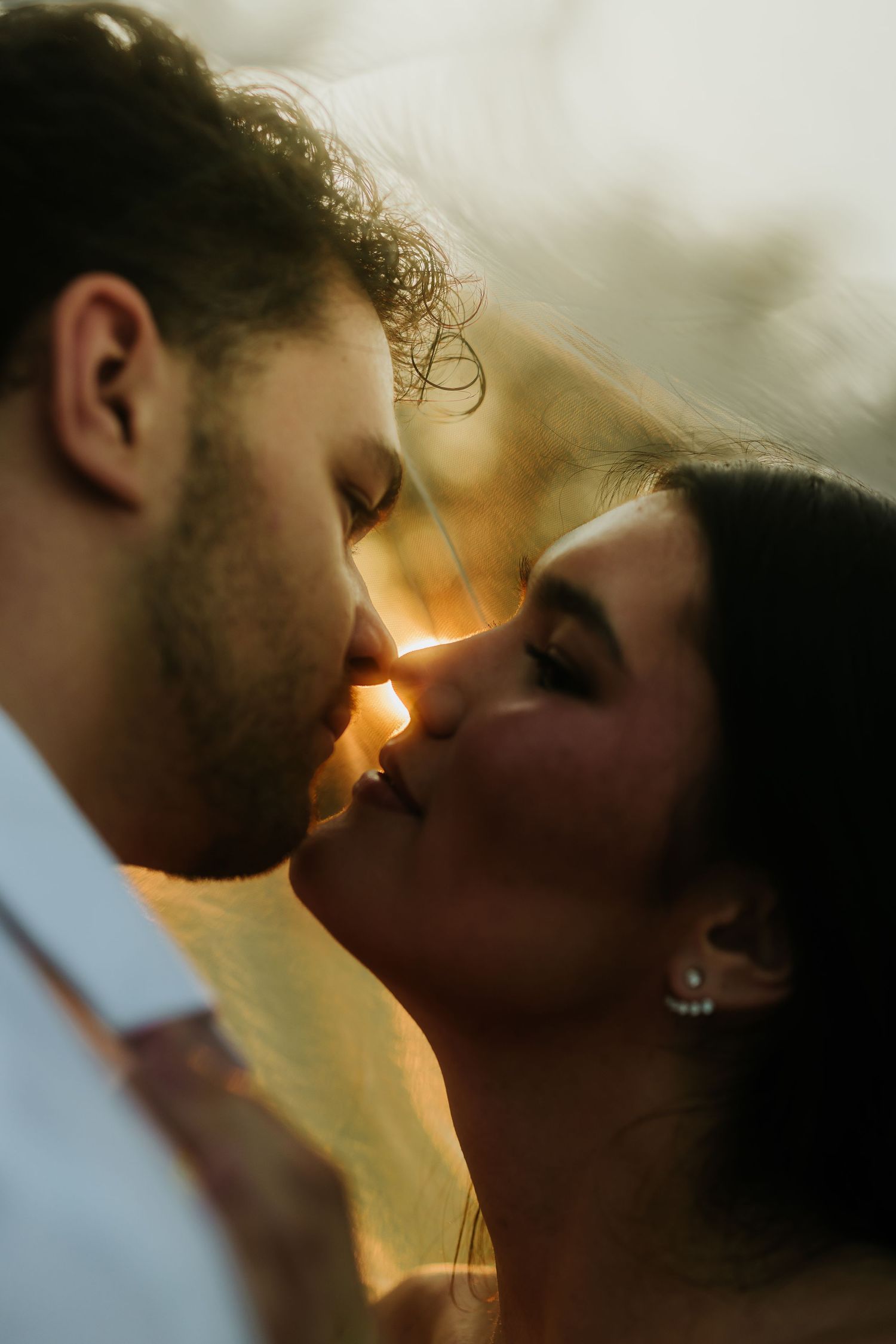 A couple shares a romantic golden hour kiss, bathed in warm sunset light, with soft bokeh background.