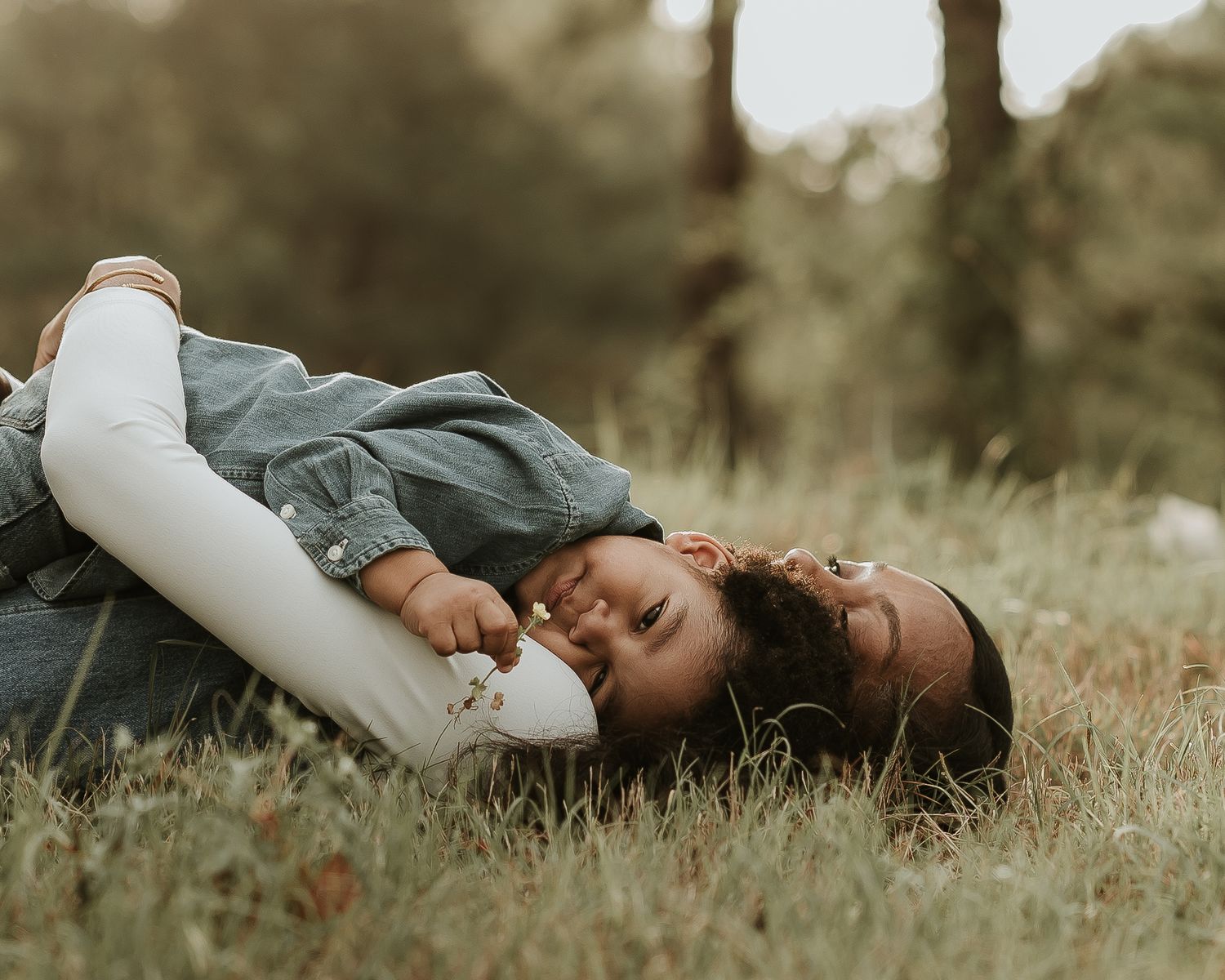 A mother and toddler lying in grass, laughing and bonding outdoors in a warm, candid family moment.