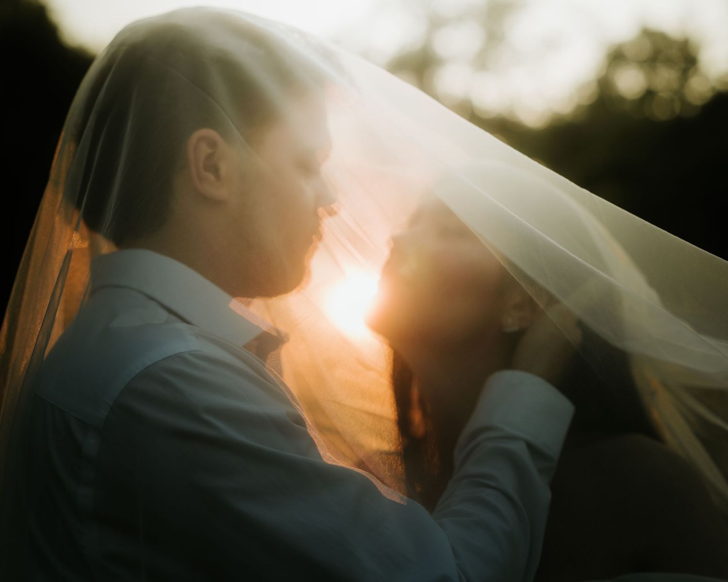 A bride and groom share a romantic kiss under a sheer wedding veil, backlit by a warm golden sunset.