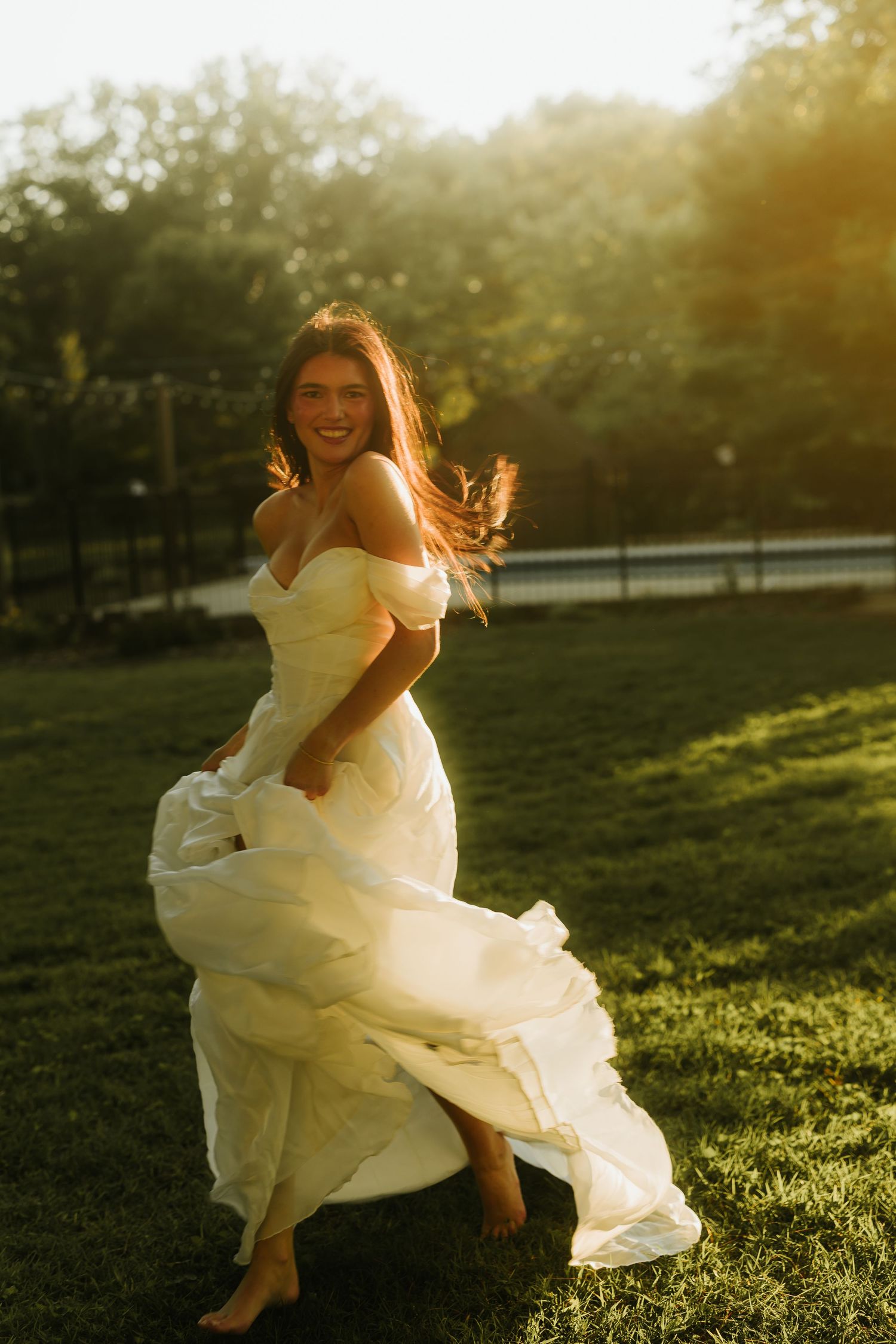 A joyful bride in an off-shoulder white gown twirls barefoot on green grass during a golden sunset.