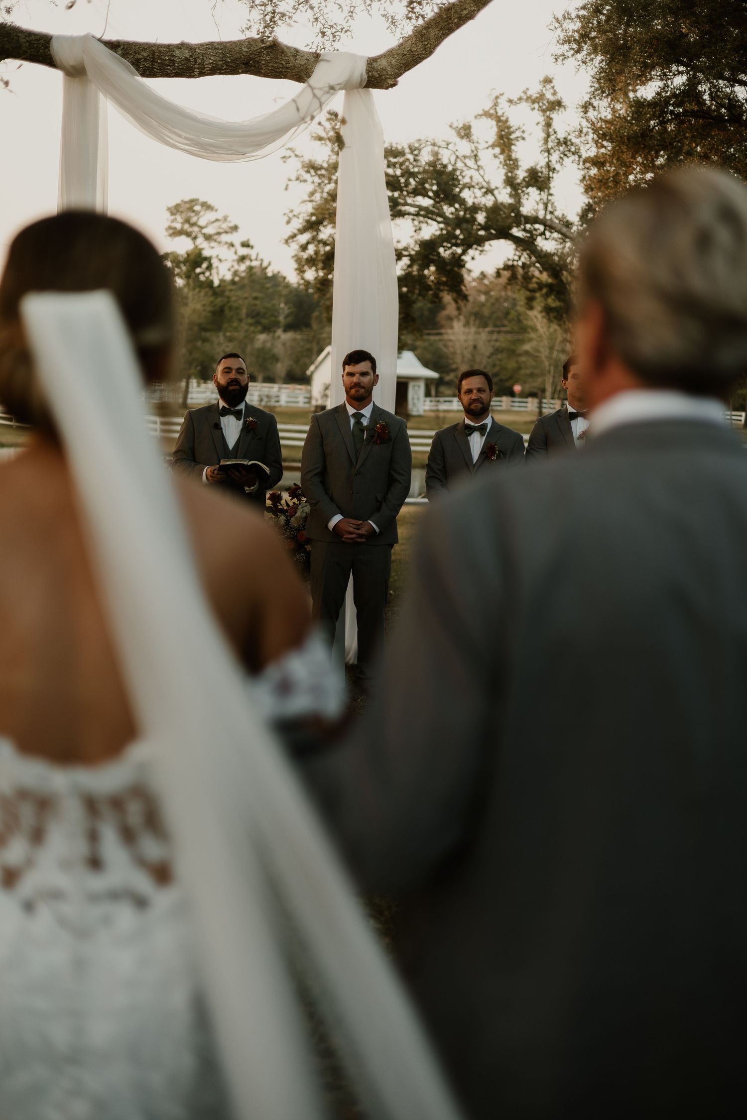 A bride walks down an outdoor aisle toward a waiting groom beneath a draped white floral arch.