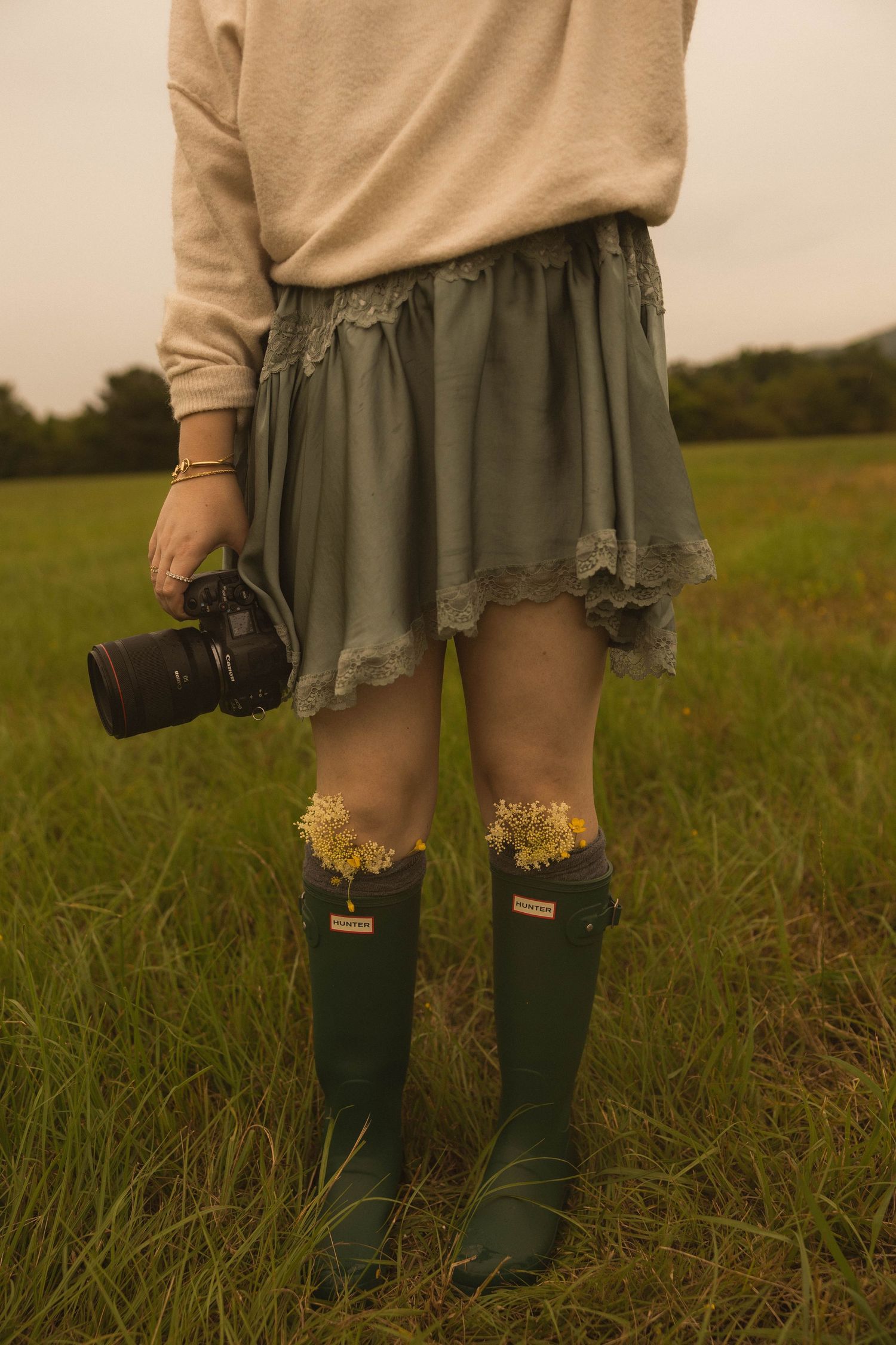 Woman in green Hunter boots and sage skirt holding a camera while standing in a grassy field.