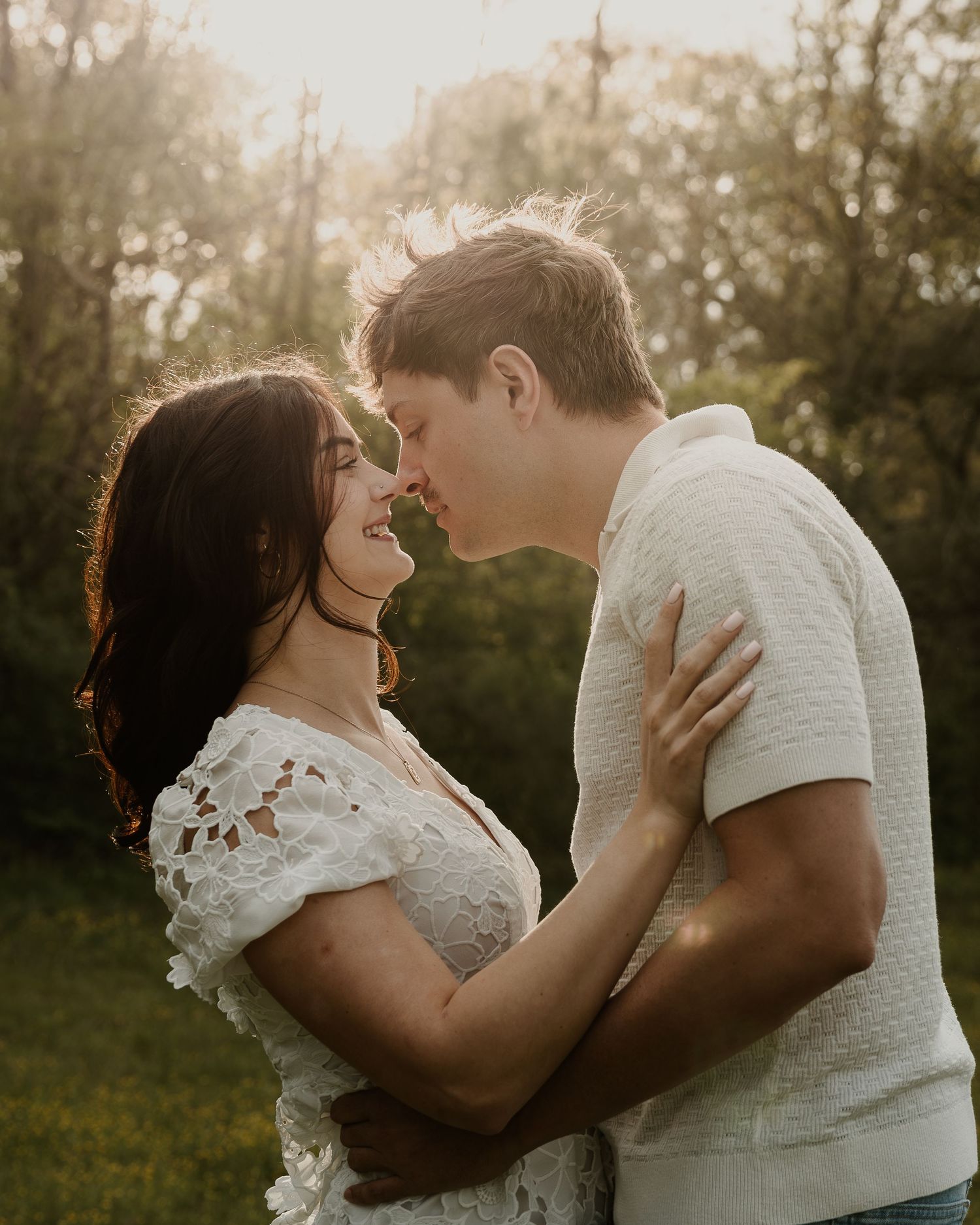 Romantic couple sharing a tender moment outdoors in golden sunset light, woman in lace dress embraced by man in white polo shirt.