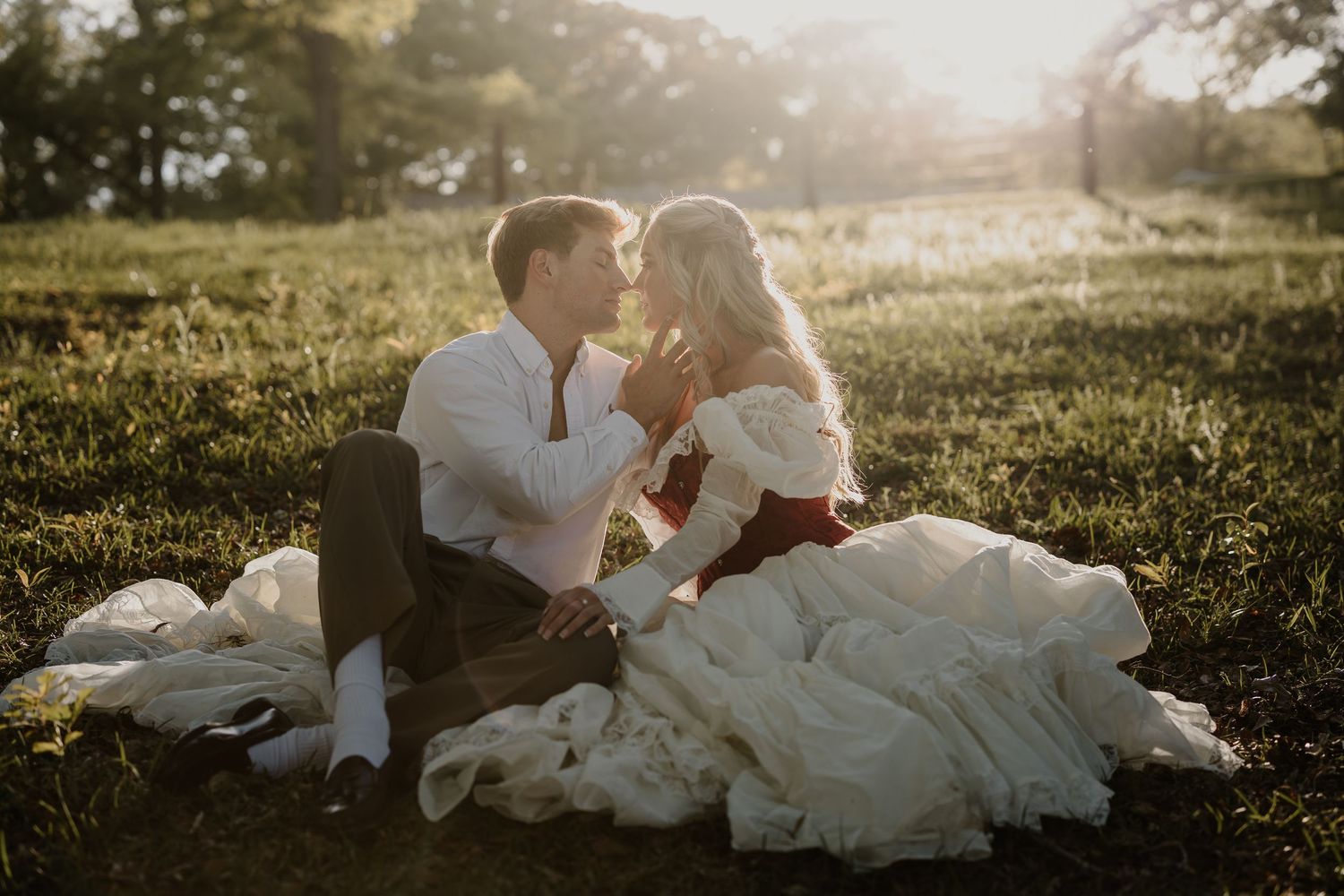 Bride in white off-shoulder gown and groom in white shirt sit on grass at golden hour, gazing lovingly at each other.