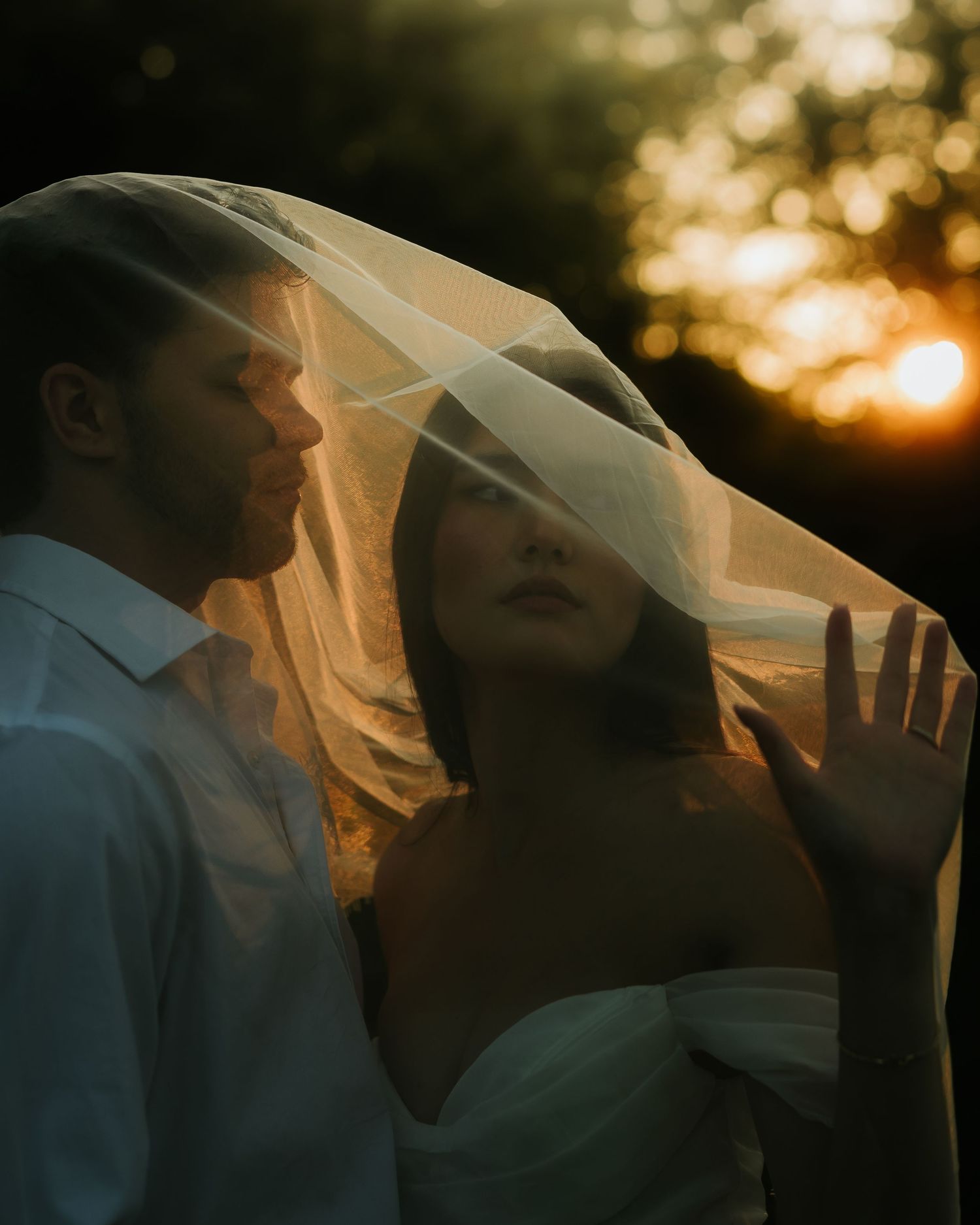 Bride and groom share romantic moment under wedding veil at golden sunset, warm bokeh background.