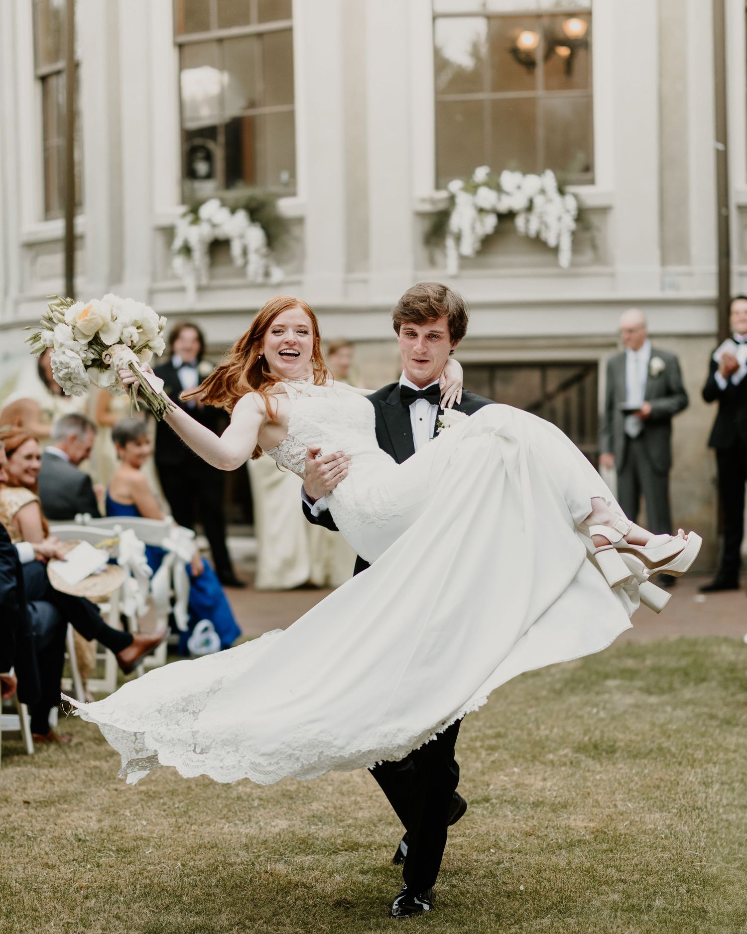 Groom carries bride in white wedding gown outdoors at elegant venue with guests seated in background.