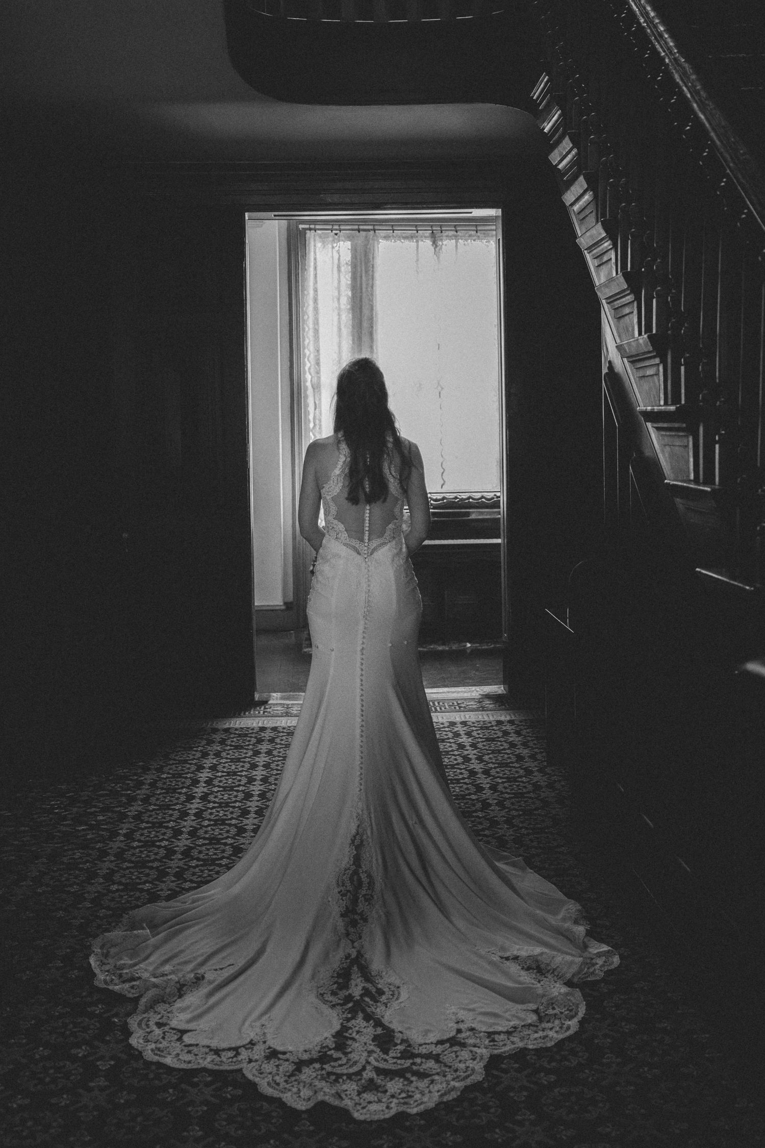 Bride in elegant lace wedding gown with long train stands before a bright window in a dark historic mansion.