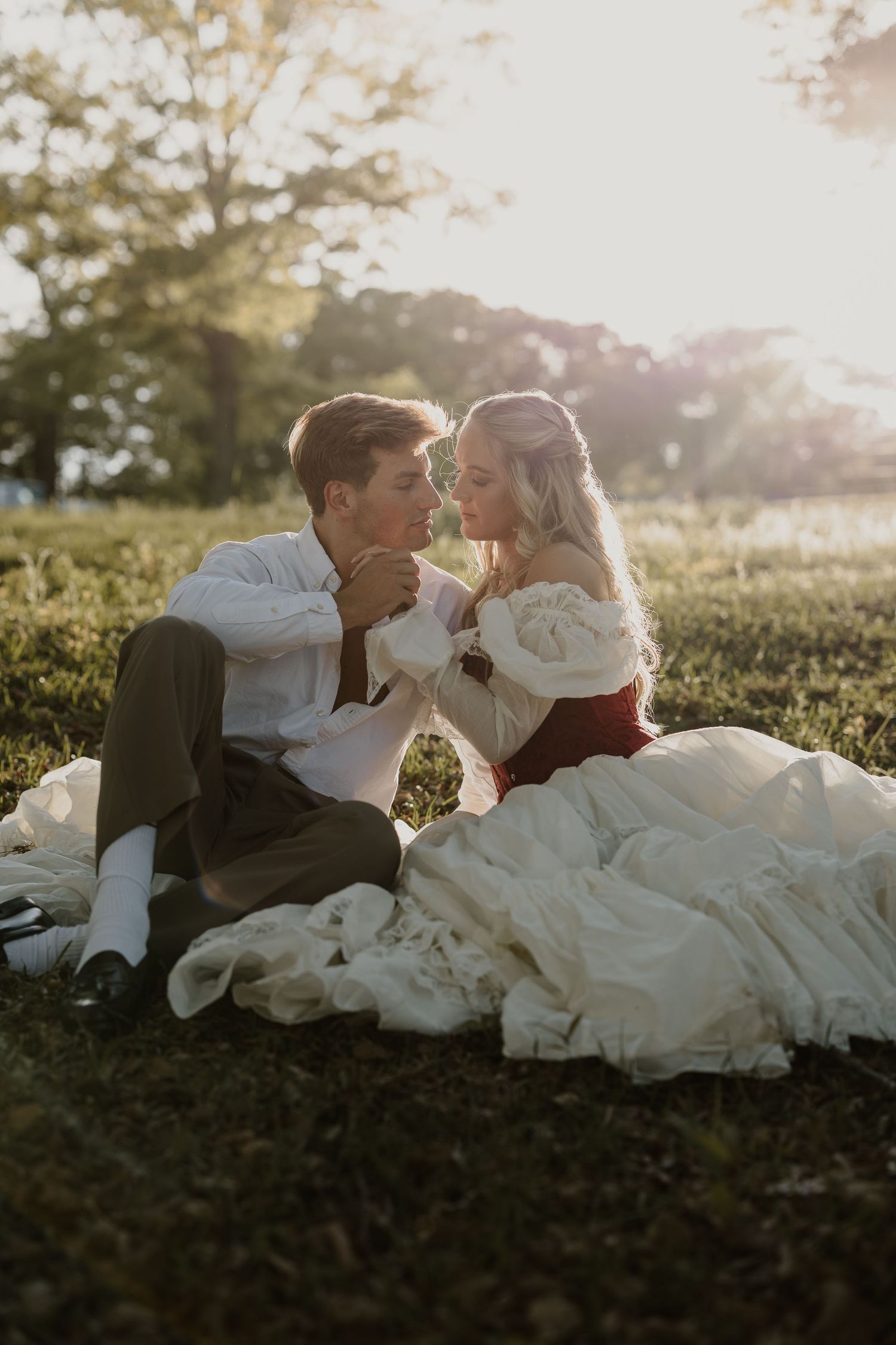 A bride in a red and white gown sits with a groom on grass, sharing a romantic moment in golden sunset light.
