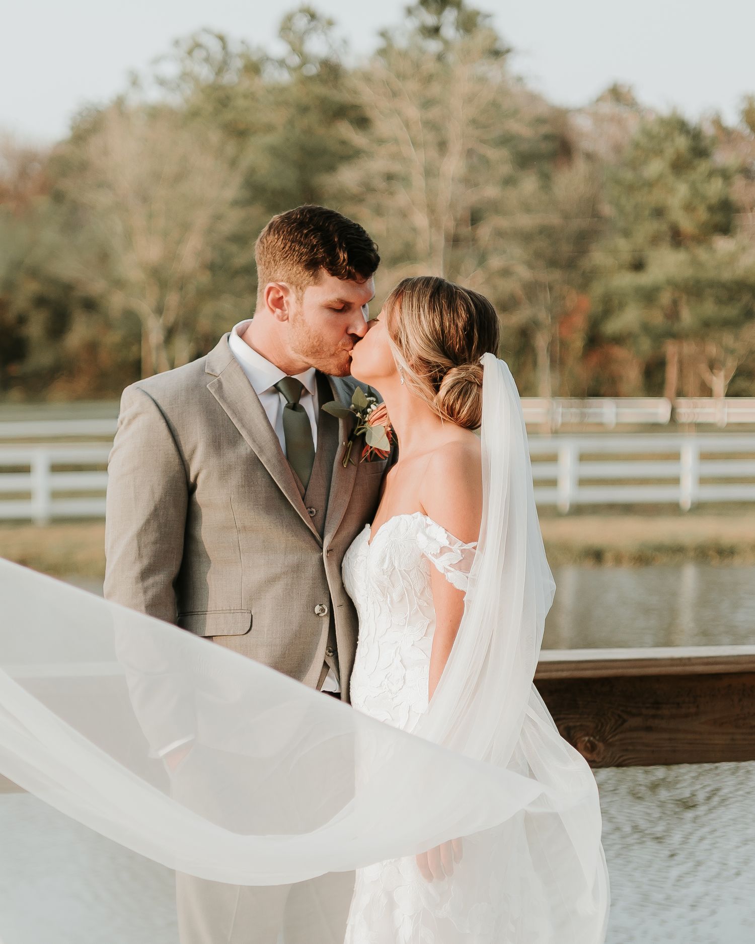 A bride and groom share a romantic kiss outdoors, with her veil flowing in the wind near a white fence and pond.