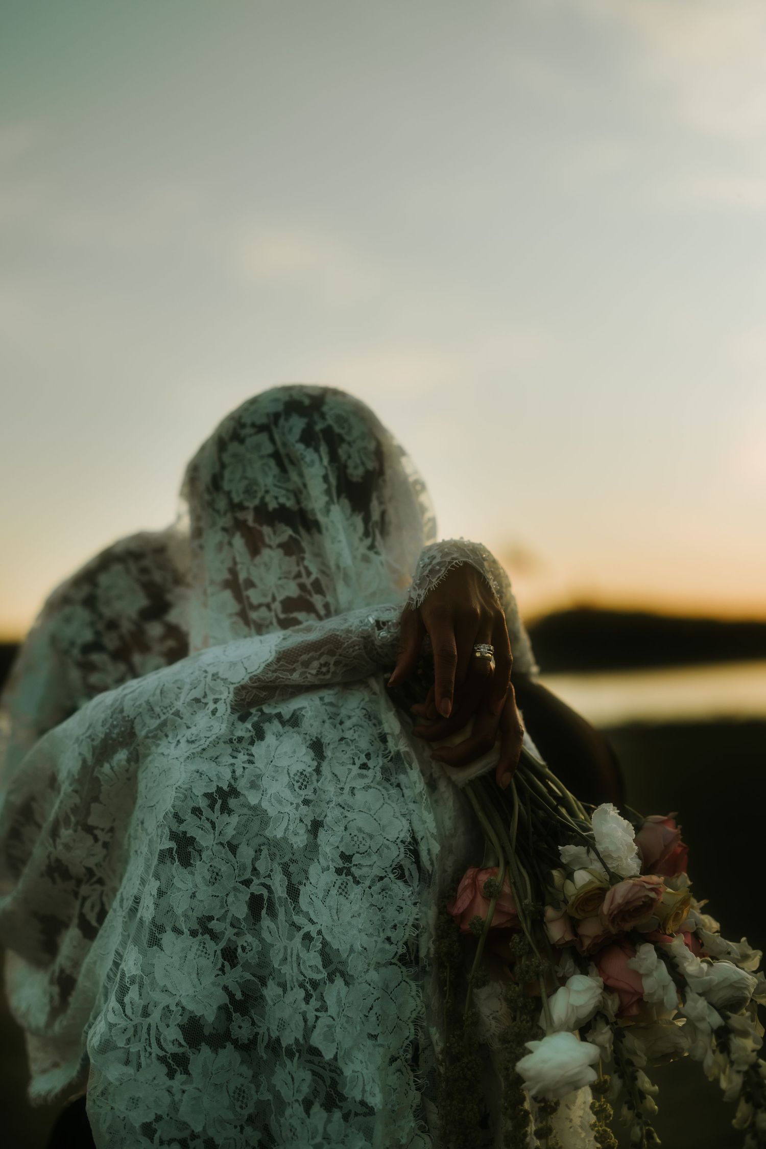 Two figures draped in white lace veils holding flowers at sunset, creating an ethereal and romantic atmosphere.