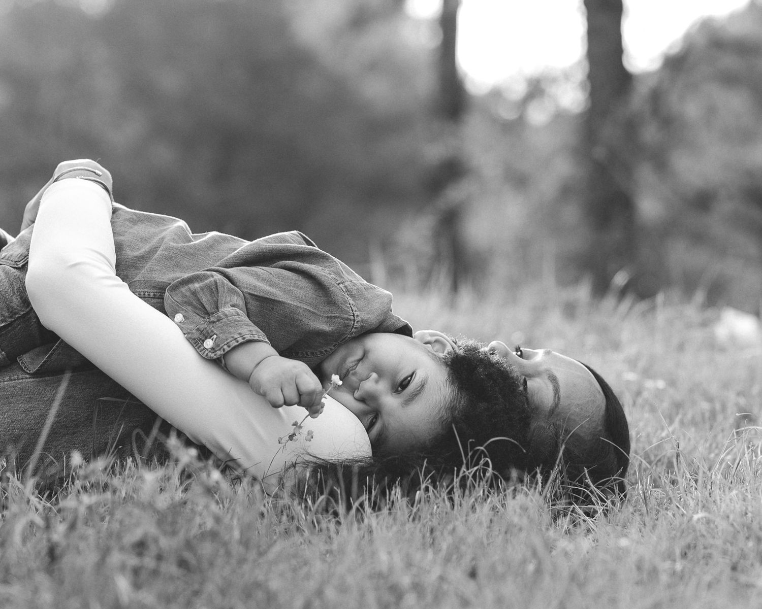 Black and white photo of a young girl lying in grass, smiling and holding a flower on a sunny day.