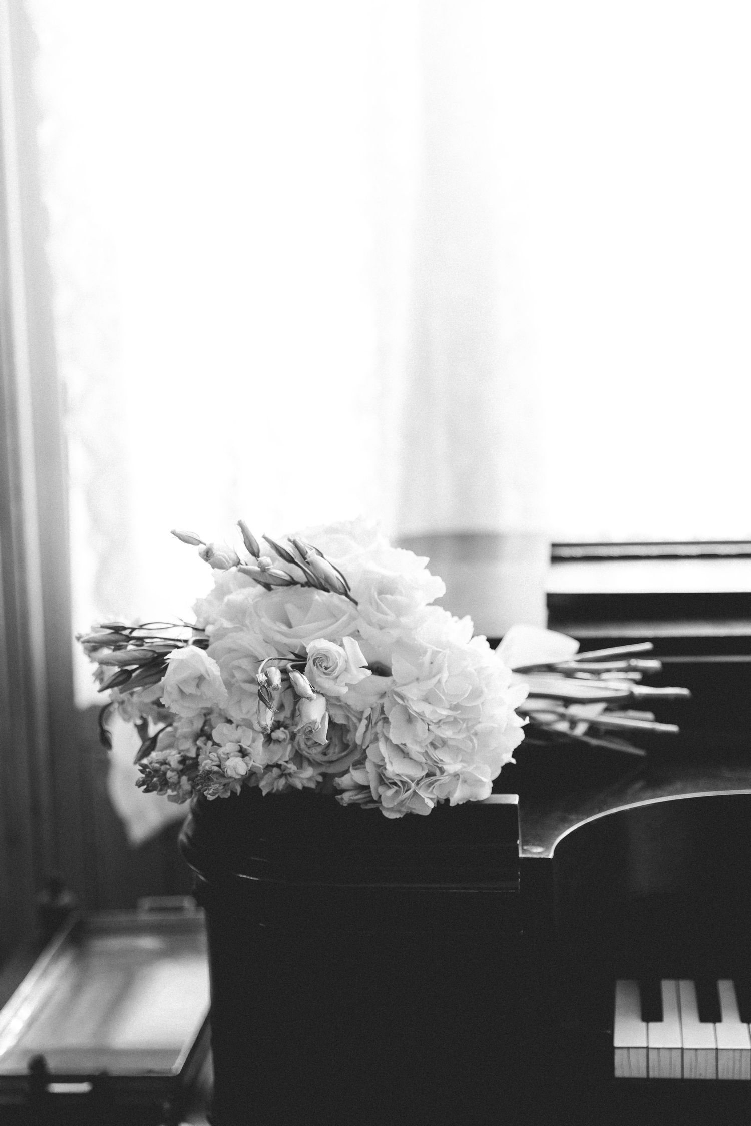 Black and white photo of a floral bouquet with roses resting on a grand piano keys near a bright window.