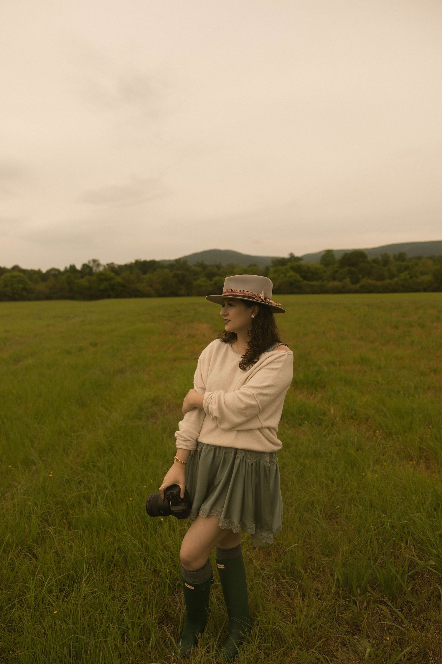 Young woman in hat, white sweater, green skirt and rain boots stands in lush field holding camera on overcast day.