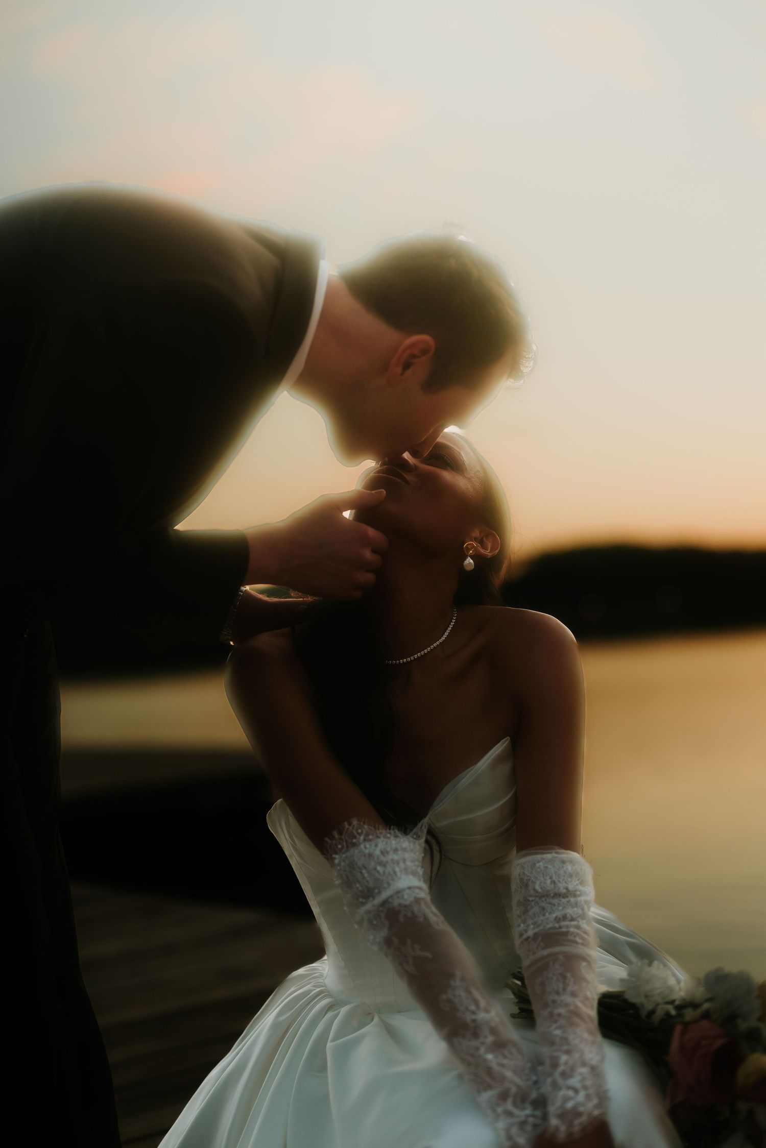 A bride in a white gown and lace gloves shares a romantic sunset kiss with her groom on a lakeside dock.