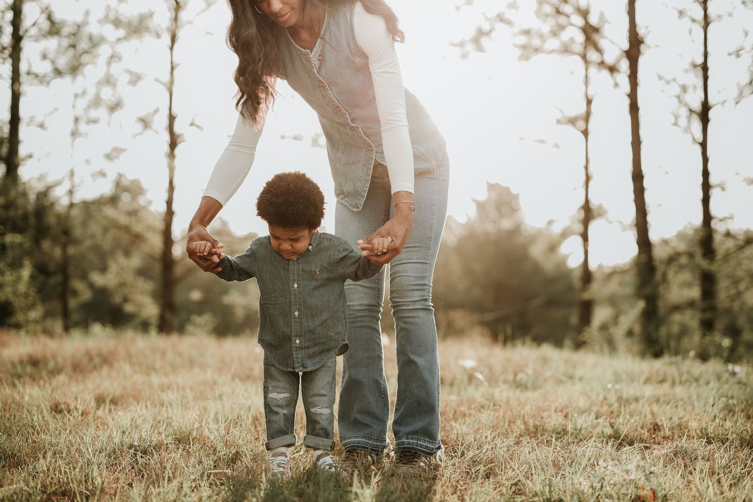 Mother helping toddler walk outdoors in sunny field, holding child's hands in warm natural light.
