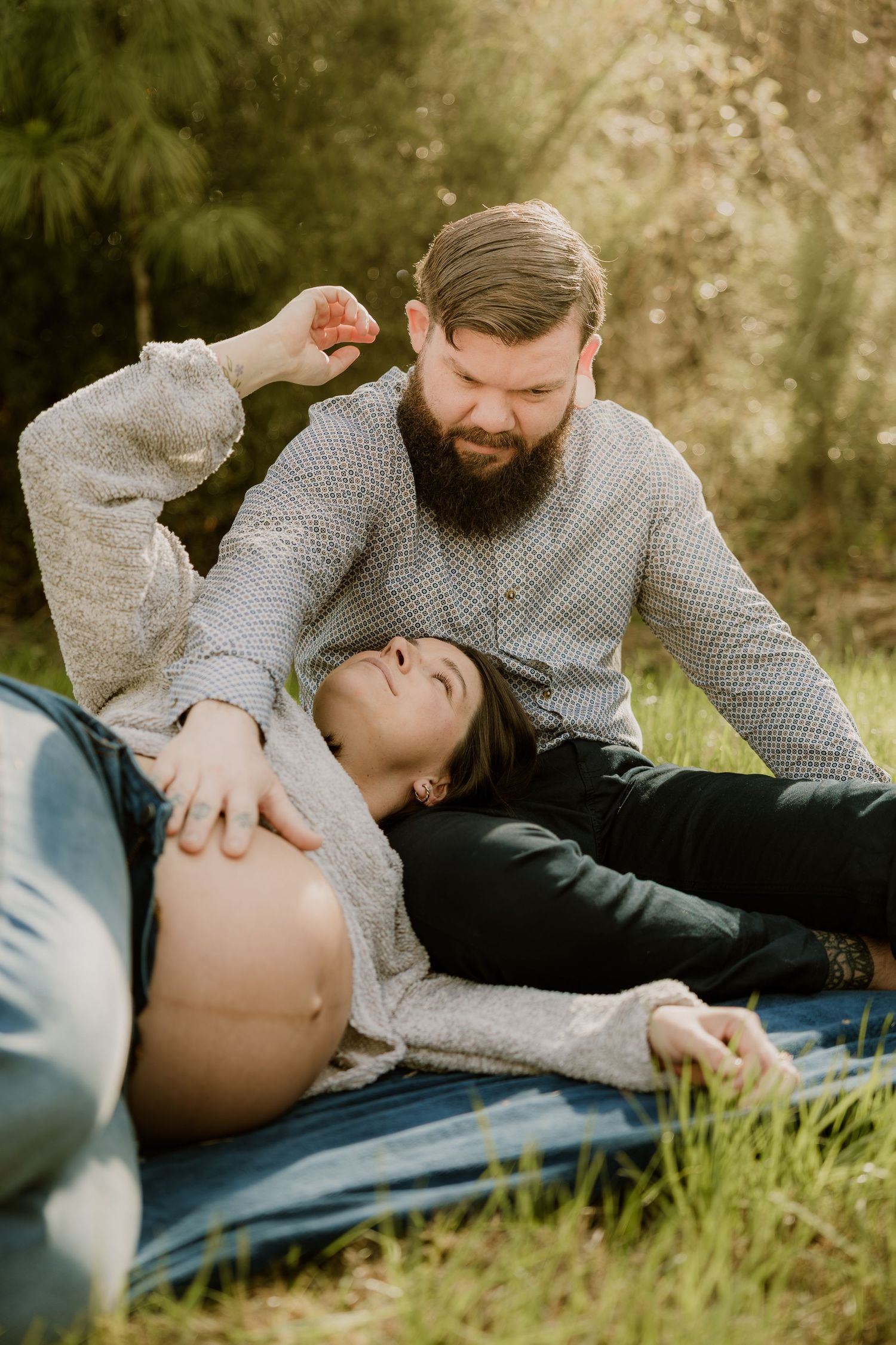 Playful maternity photo of couple relaxing on blanket in sunlit garden.
