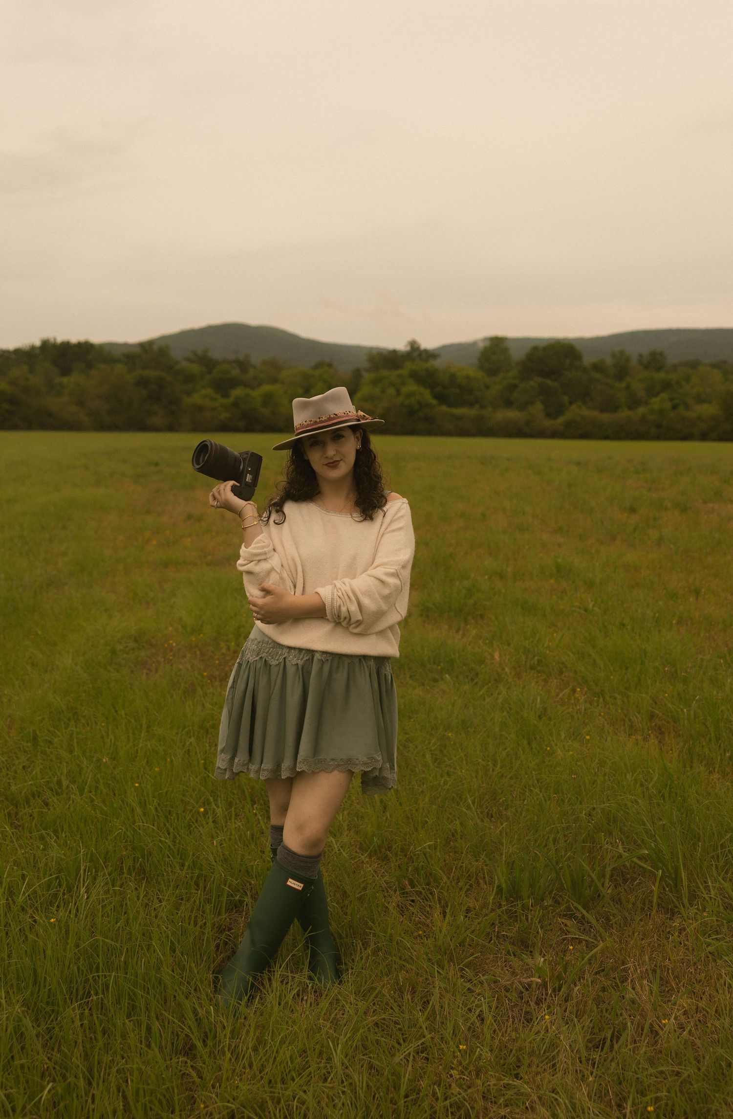 Young woman in cream sweater, green skirt, boots, and hat holds camera in lush green field with mountains behind.