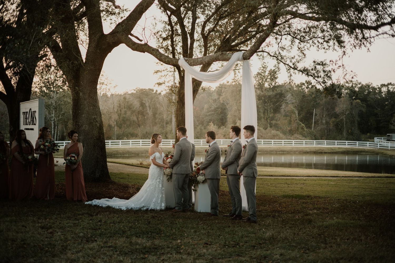 Outdoor wedding ceremony under a large oak tree with wedding party in gray suits and burgundy dresses at a rustic venue.