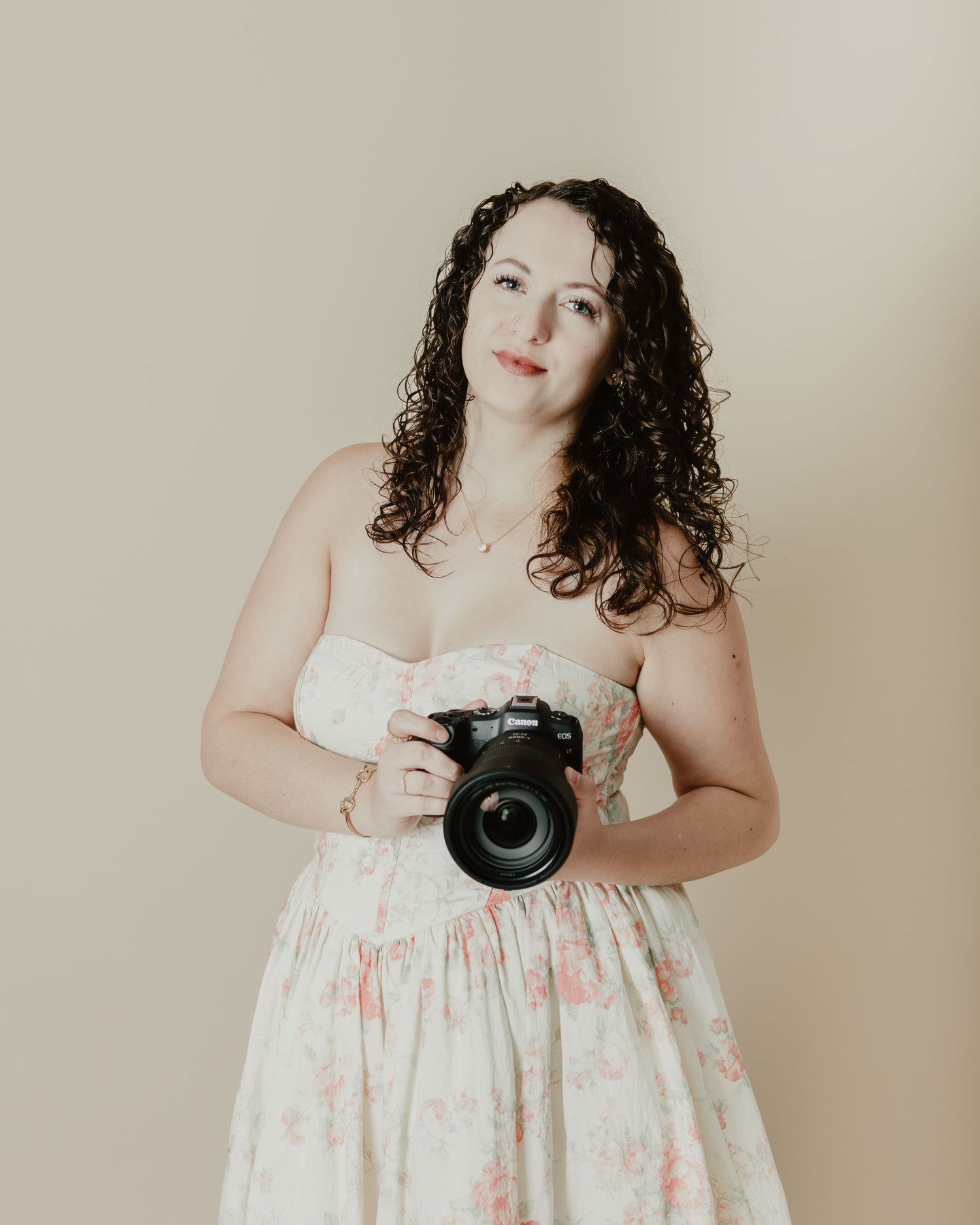 A woman with curly dark hair holds a DSLR camera, wearing a floral strapless dress against a beige background.