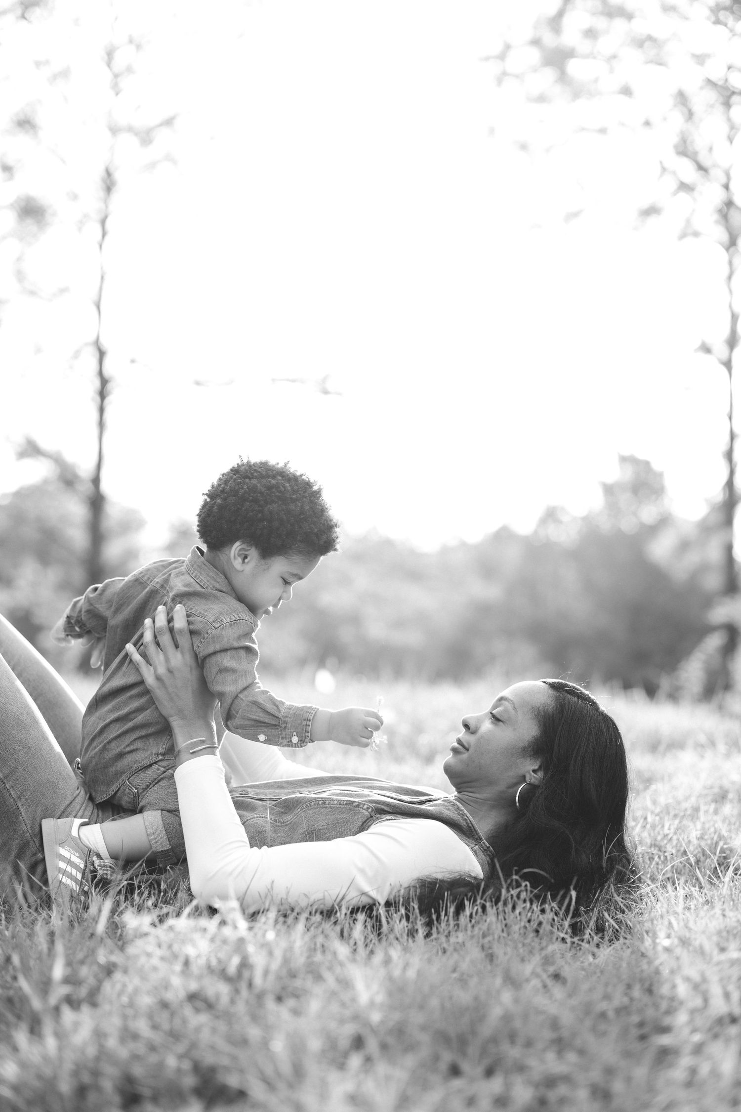 Black and white photo of a mother lying on grass lovingly holding her baby in a sunny park.