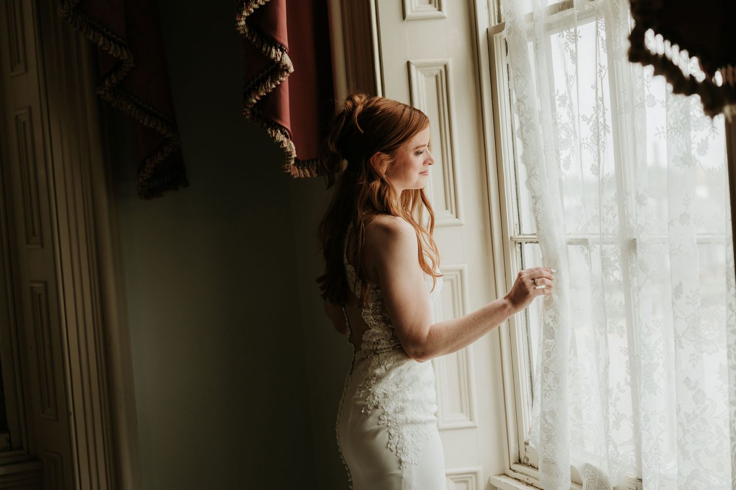 Bride in white lace gown gazing out a sunlit window, holding sheer curtain in elegant wedding portrait.
